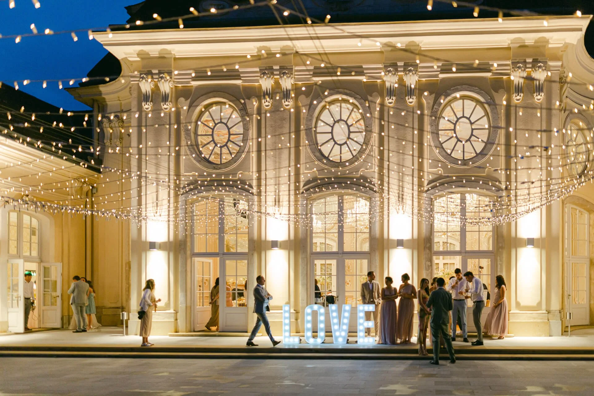 Abendszene vor einem beleuchteten Gebäude mit dekorativen Lichterketten. Eine Gruppe von Menschen in eleganter Kleidung steht vor dem Gebäude. Im Vordergrund leuchten die Buchstaben "LOVE" in großen, hellen Lettern.