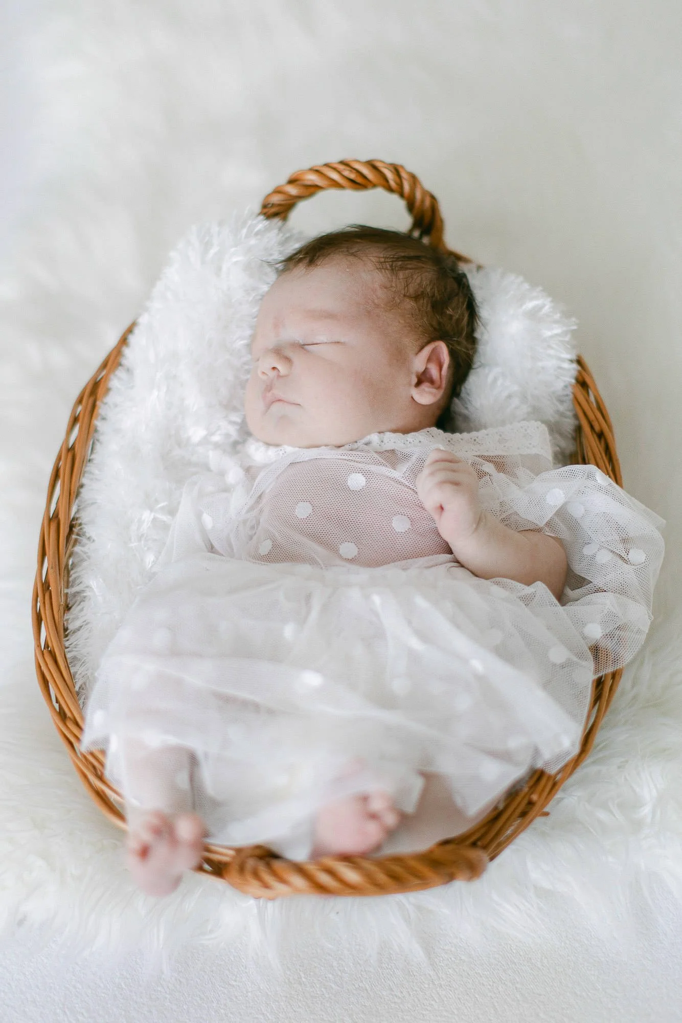 Schlafendes Baby in weißem Kleid in Weidenkorb auf Fellunterlage.