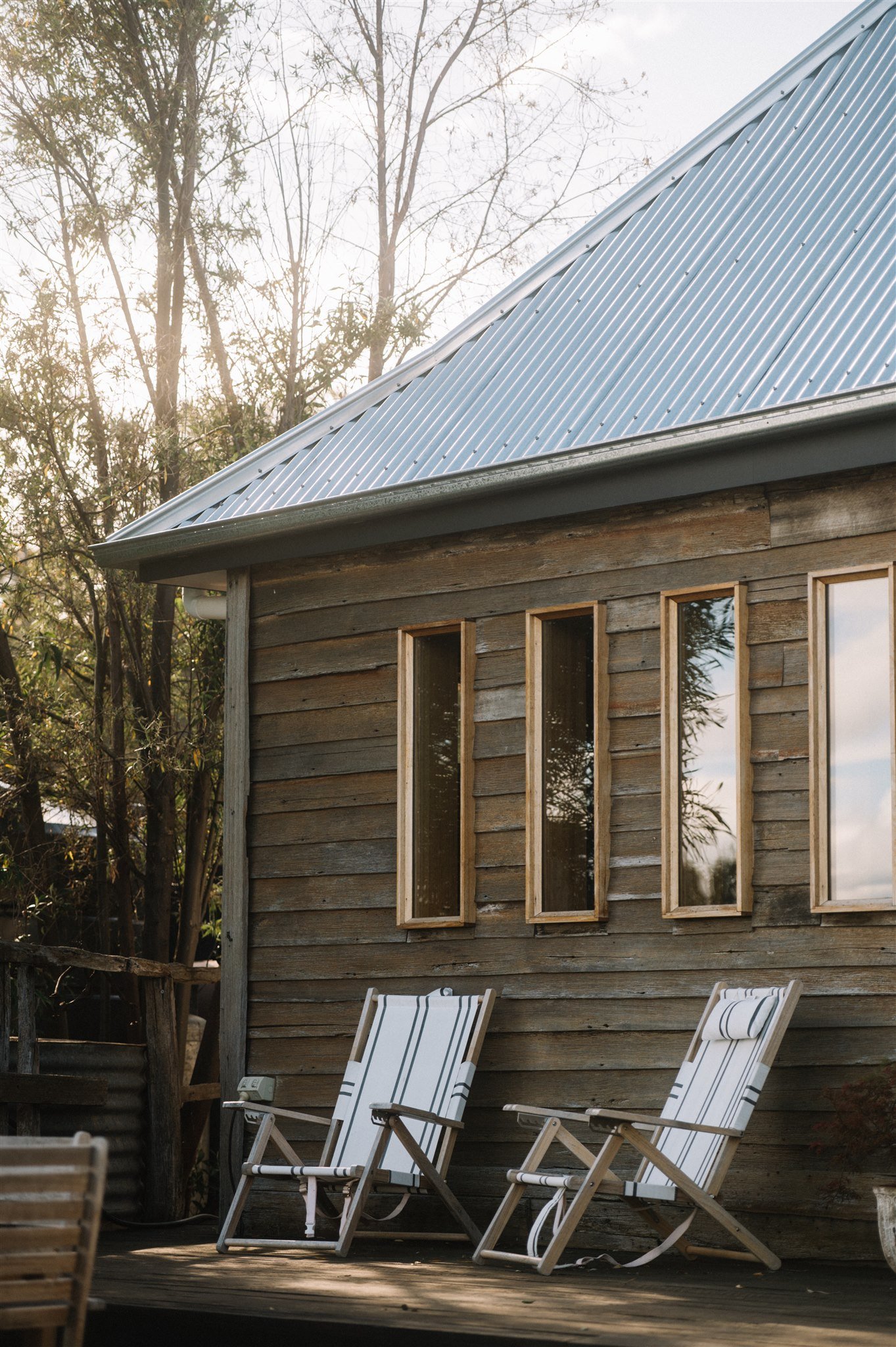 Outdoor scene with a wooden house having tall narrow windows, and two striped patio chairs on a wooden deck, surrounded by trees.