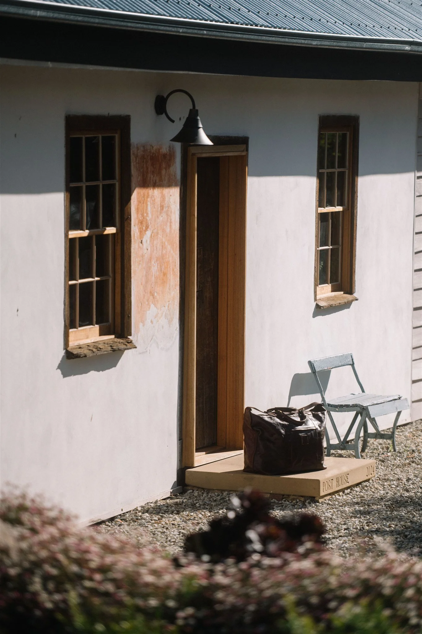 Exterior of a small, white building with two wooden windows, a black outdoor wall light, a small concrete porch with a brown bag and a white bench.