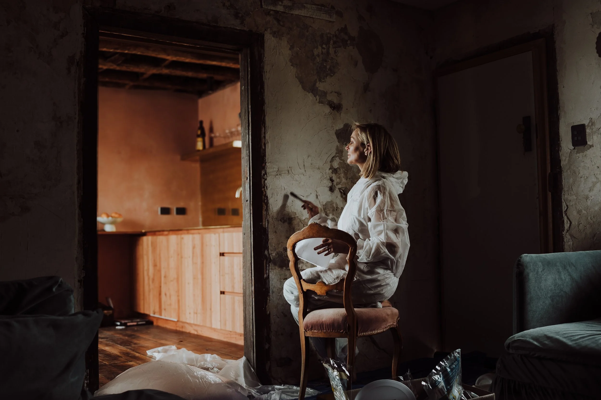 A woman in white pajamas sitting on a vintage chair, holding a mug, and looking thoughtfully out of a doorway into a kitchen with wooden cabinets.