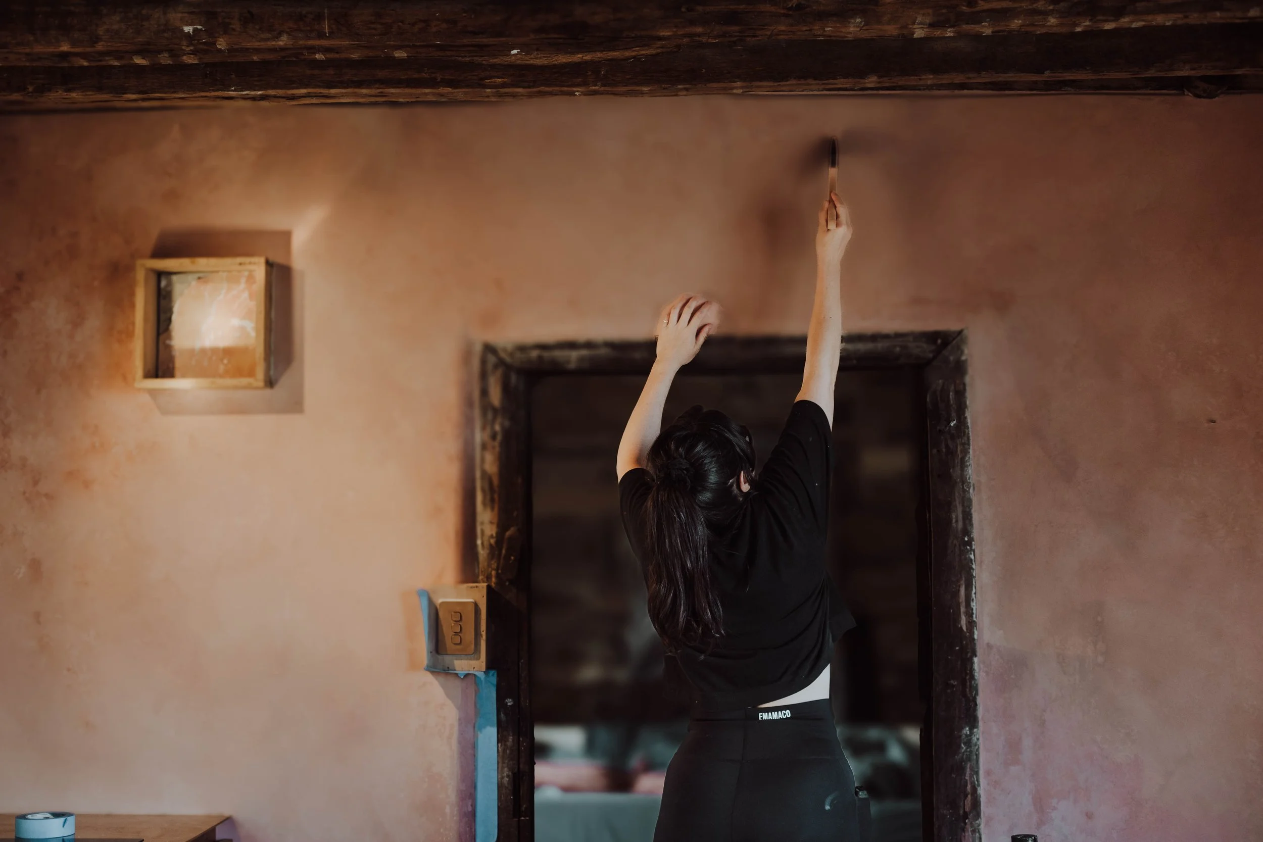 A woman in black clothing reaching up to a ceiling with her right arm, her back turned to the camera, in a room with a pinkish wall and wooden trim.