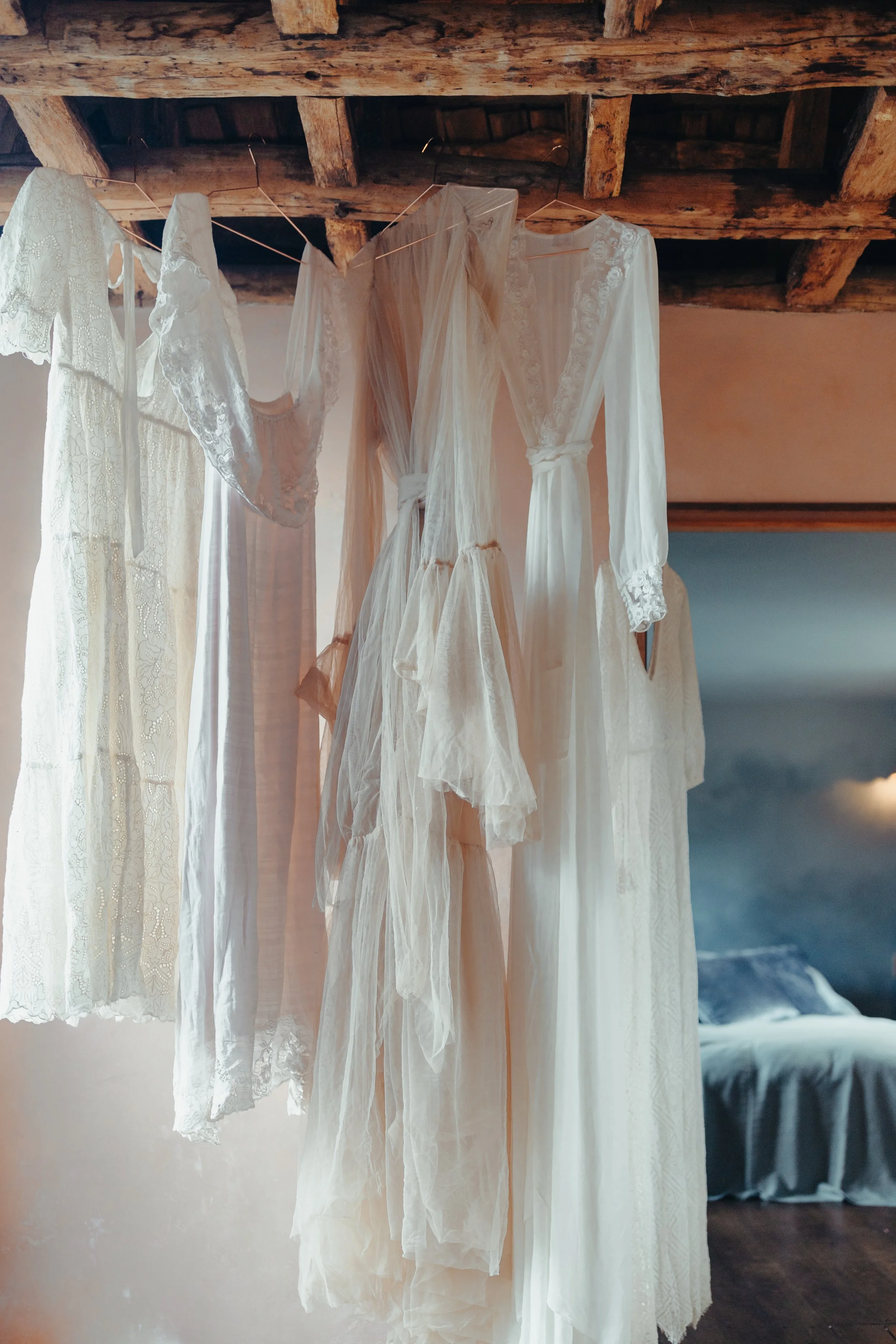 Several white and cream-colored garments, including dresses and robes, hanging on a line indoors with a rustic wooden ceiling.