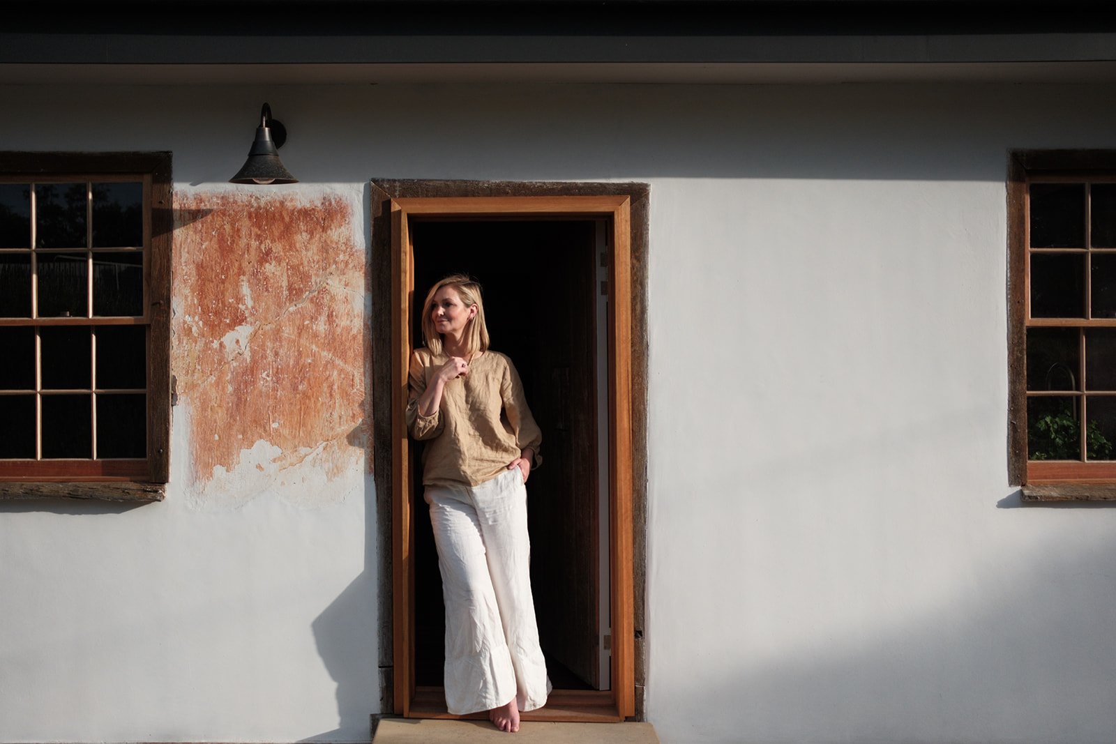 A woman standing barefoot in the doorway of a house, looking outside with her hand on her chest and her other hand in her pocket. The house has white walls, wooden window frames, and a partially distressed wall near the window.