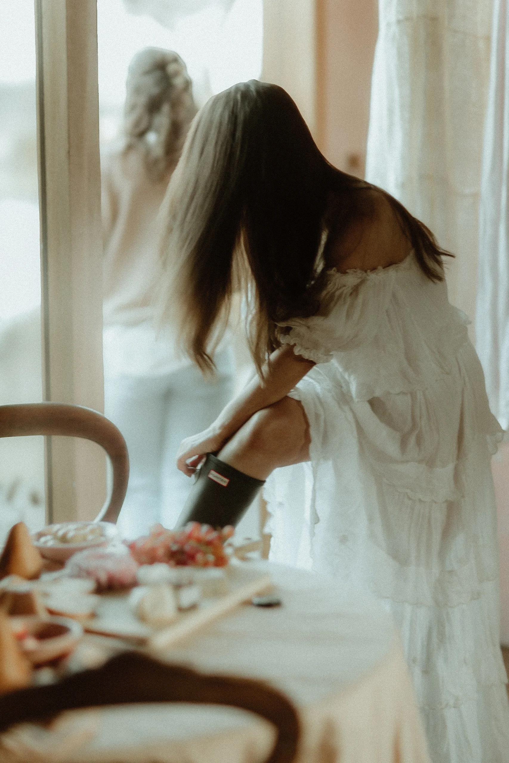 A woman in a white dress using a meat grinder on a table with food, with a doll in the background outside a window.