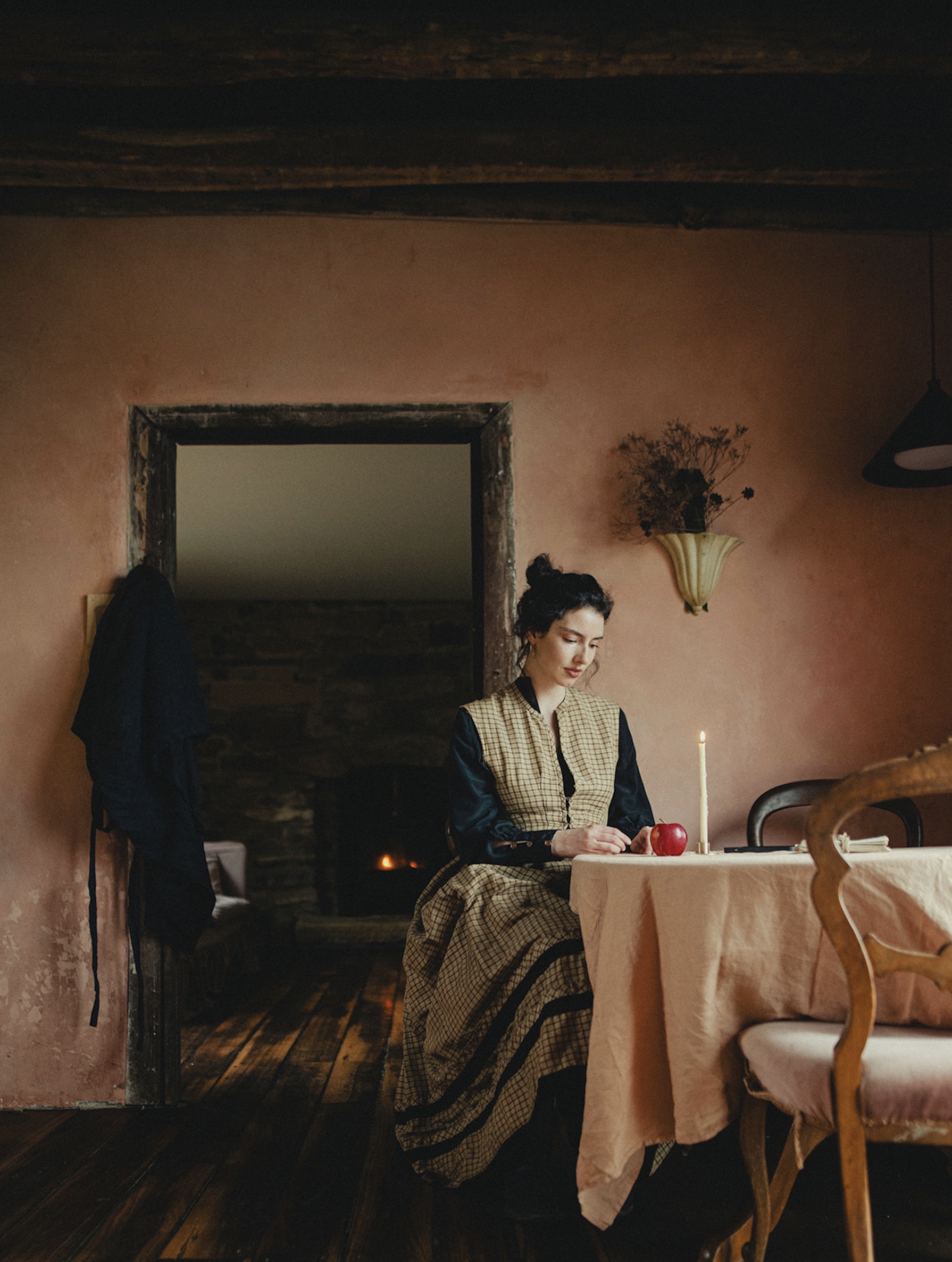 Woman sitting at a dining table with a candle and an apple in a rustic room with pink walls and a stone fireplace.