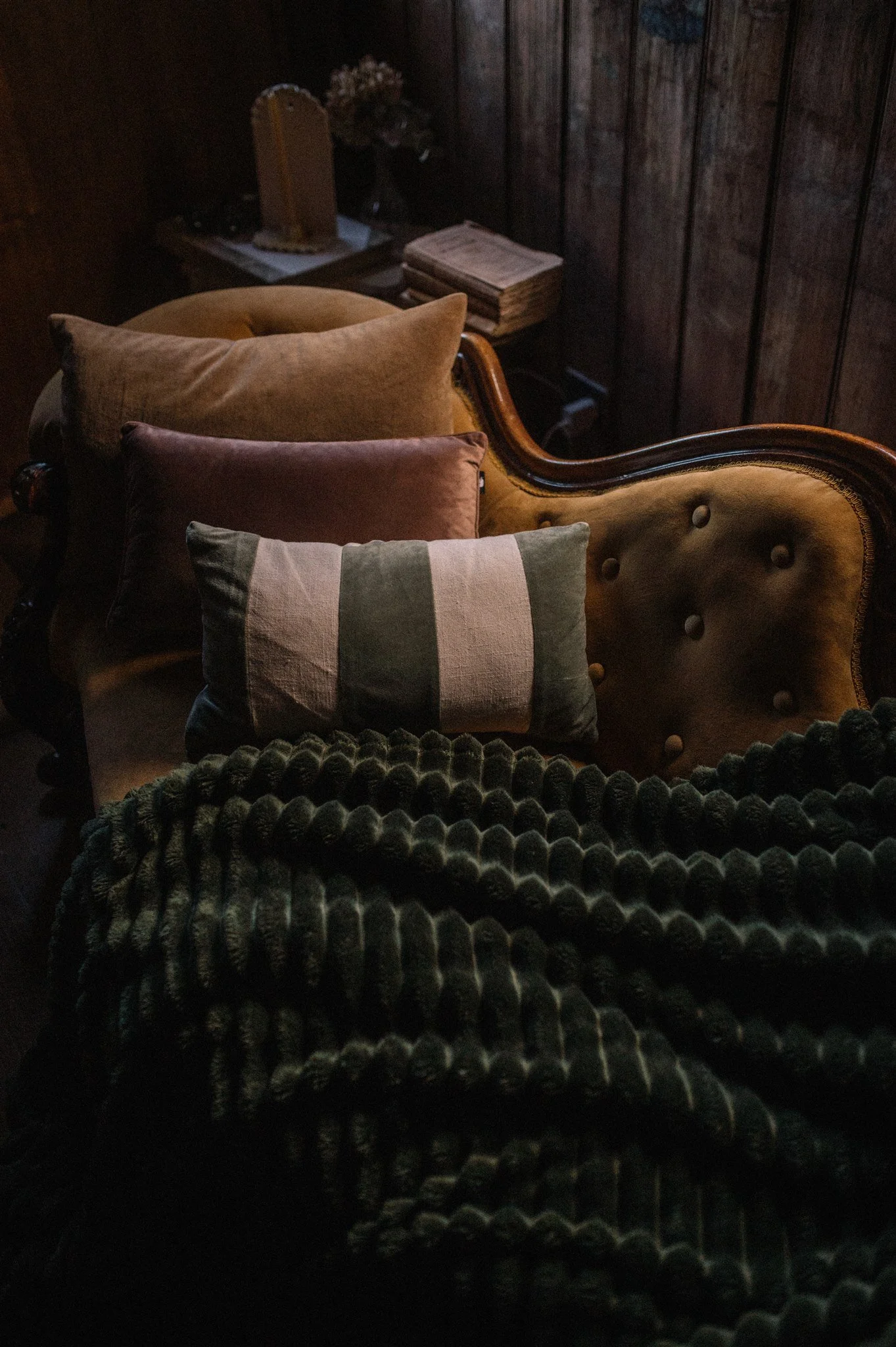 A cozy, vintage-style living room corner with a tufted, mustard-colored sofa decorated with three pillows and a plush, textured dark blanket. There is a small wooden side table with stacked books and a decorative object against a dark wooden wall.