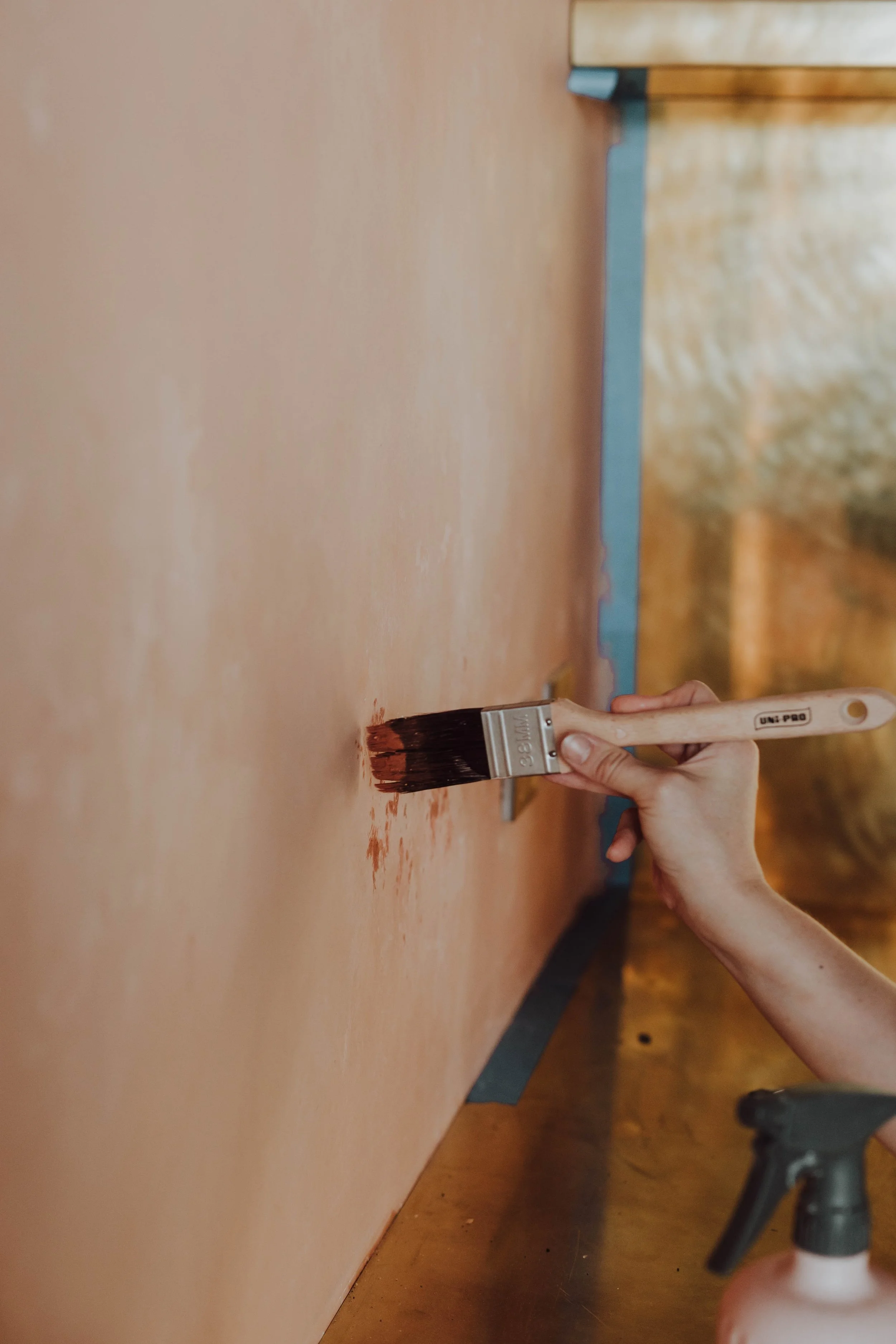 Person painting a wall with a brown paintbrush, applying brown paint to a peach-colored wall.
