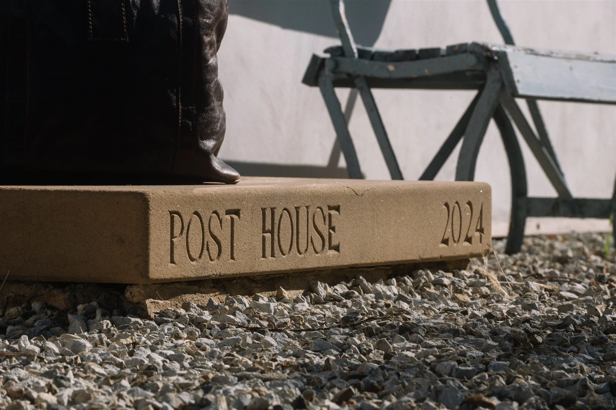 Close-up of a concrete base with engraved text 'POST HOUSE 2024'; there is a leather bag partially visible on top of the base, and a weathered wooden bench in the background on gravel ground.