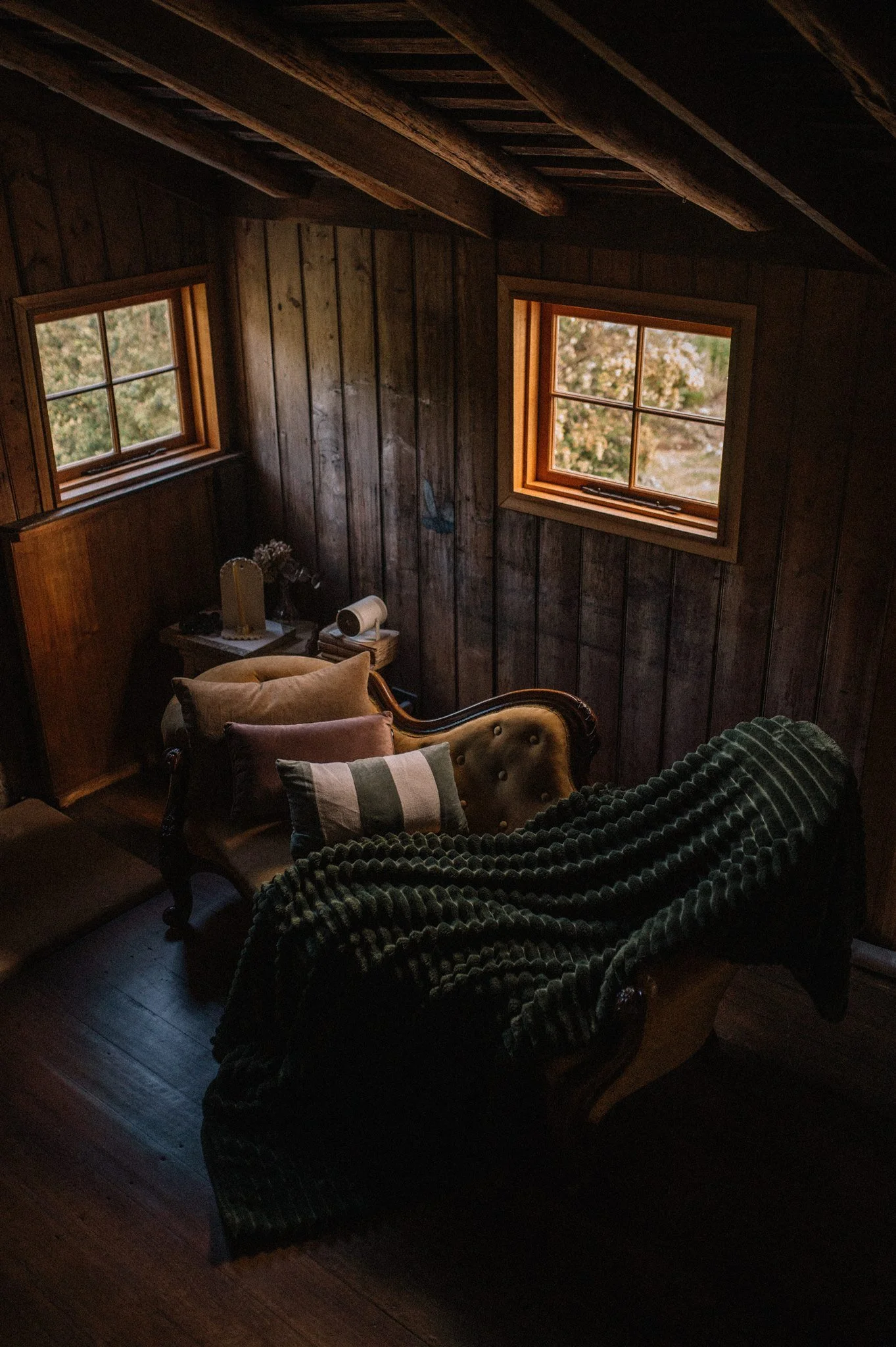 A cozy living room with wooden walls and two small windows, a vintage sofa with cushions and a blanket, and a small side table with decorative items.