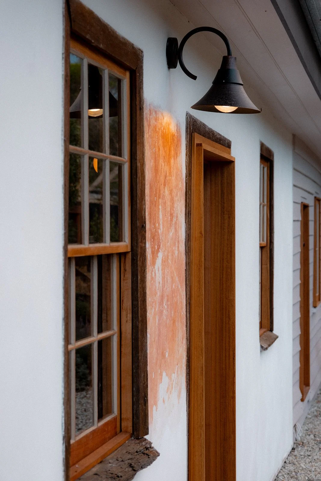 Close-up of a rustic exterior wall with wooden window frames, a wall-mounted black metal sconce with a downward-facing lamp, and a partially painted wall section in a barn-style building.