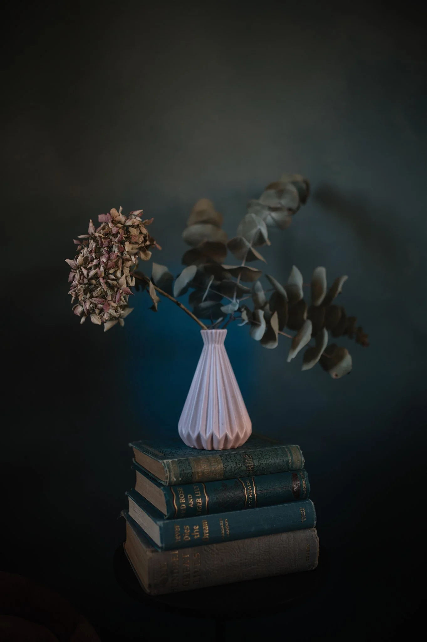 A still life arrangement with a pink pleated vase holding dried flowers and branches, placed on a stack of old hardcover books against a dark background.