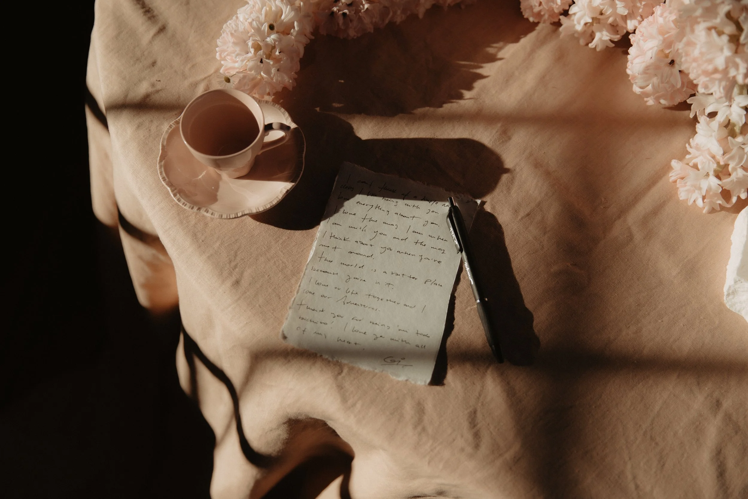 A peach tablecloth drapes a round table, flowers in the upper right corner and a peachy pink coffee up in the left.  A piece of hand torn paper filled with words  and a pen nearby.