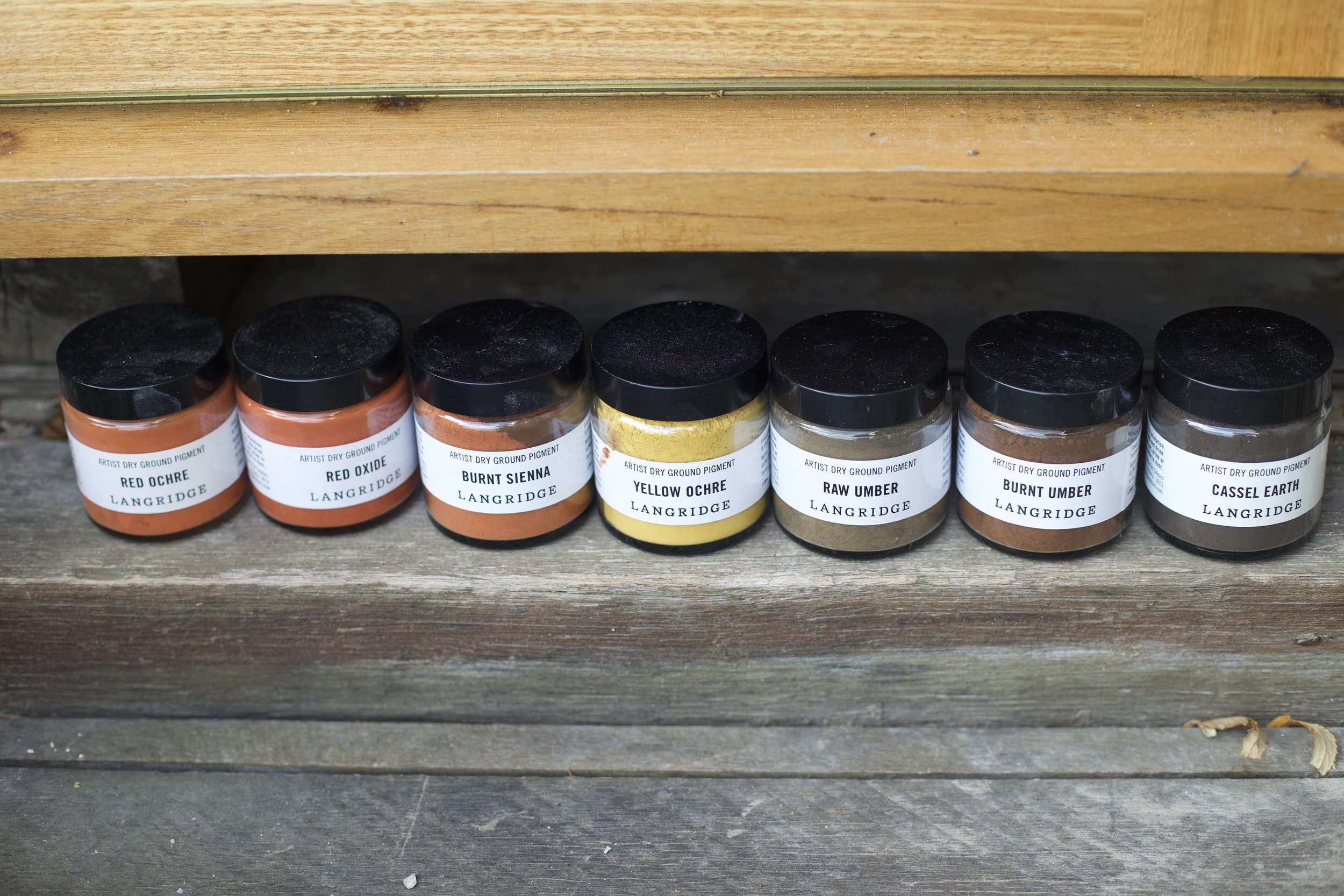 Six jars of pigment labeled from left to right: Red Ochre, Red Oxide, Burnt Sienna, Yellow Ochre, Raw Umber, Burnt Umber, and Cassel Earth, on a wooden surface with a wooden backdrop.