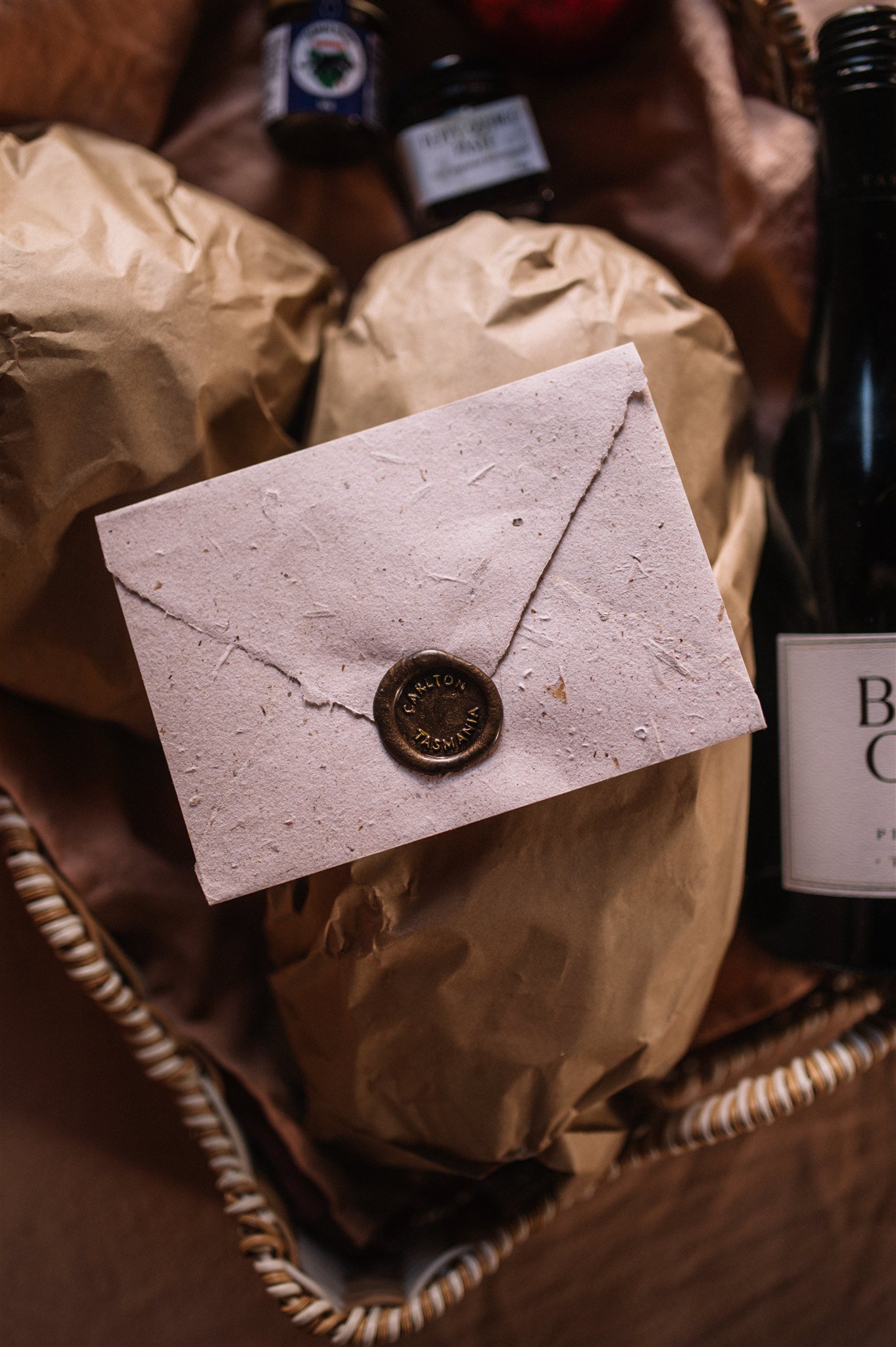 A white textured envelope with a bronze wax seal, placed on top of brown paper packages inside a woven basket.