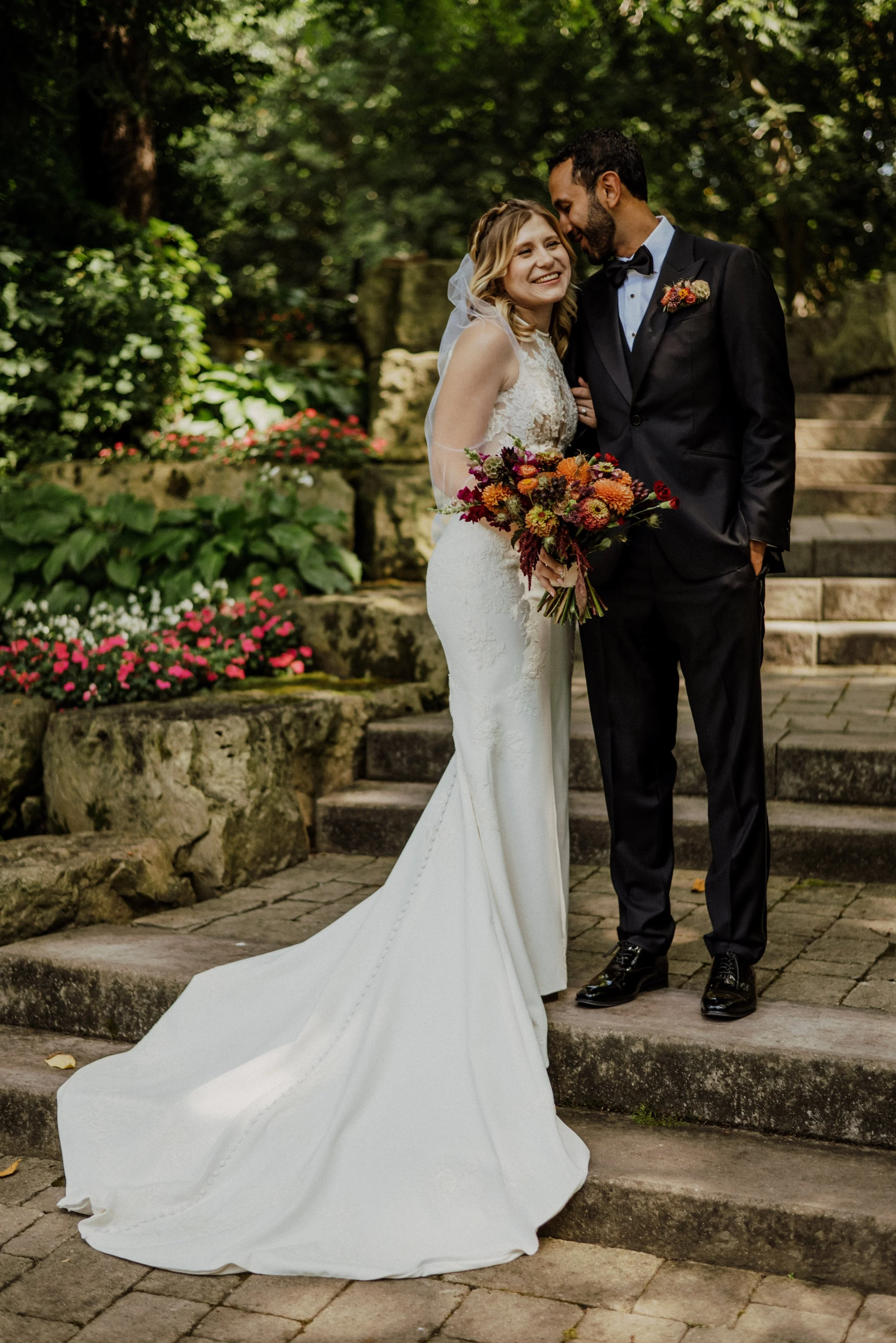 A bride and groom smiling and standing closely together outdoors on stone steps, with lush green foliage and flowers in the background. The bride is holding a bouquet of flowers and is dressed in a lace wedding gown with a long train, while the groom is in a black tuxedo with a boutonniere.