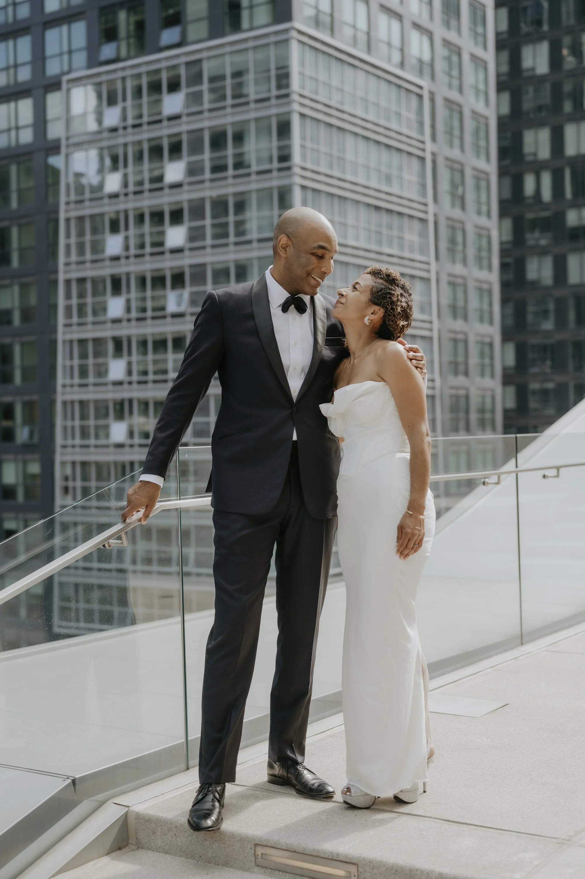 A smiling couple in wedding attire sharing an intimate moment on a city balcony with modern glass buildings in the background.