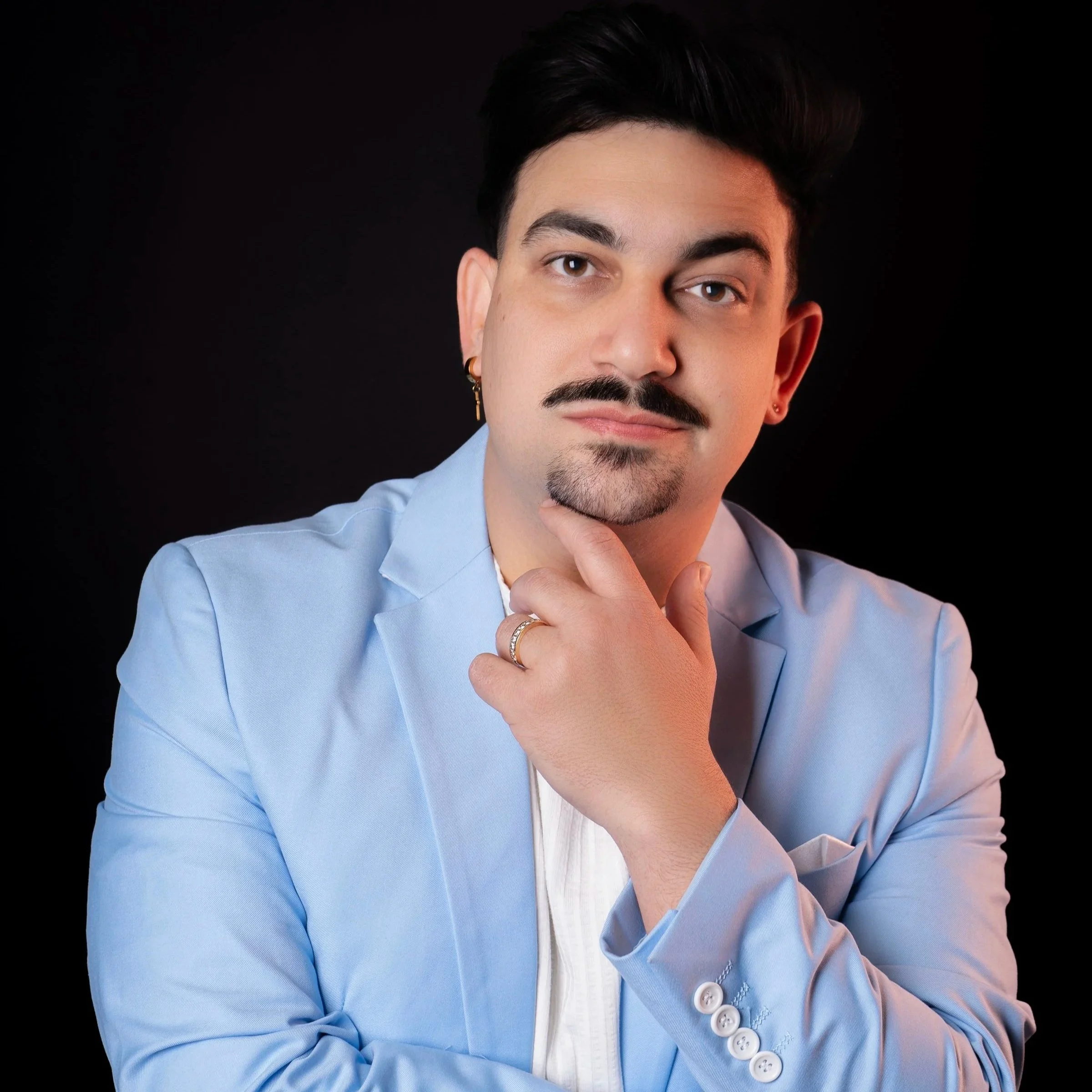 Portrait of a young man with dark hair, mustache, and goatee, wearing a light blue blazer, with his hand on his chin, against a black background.