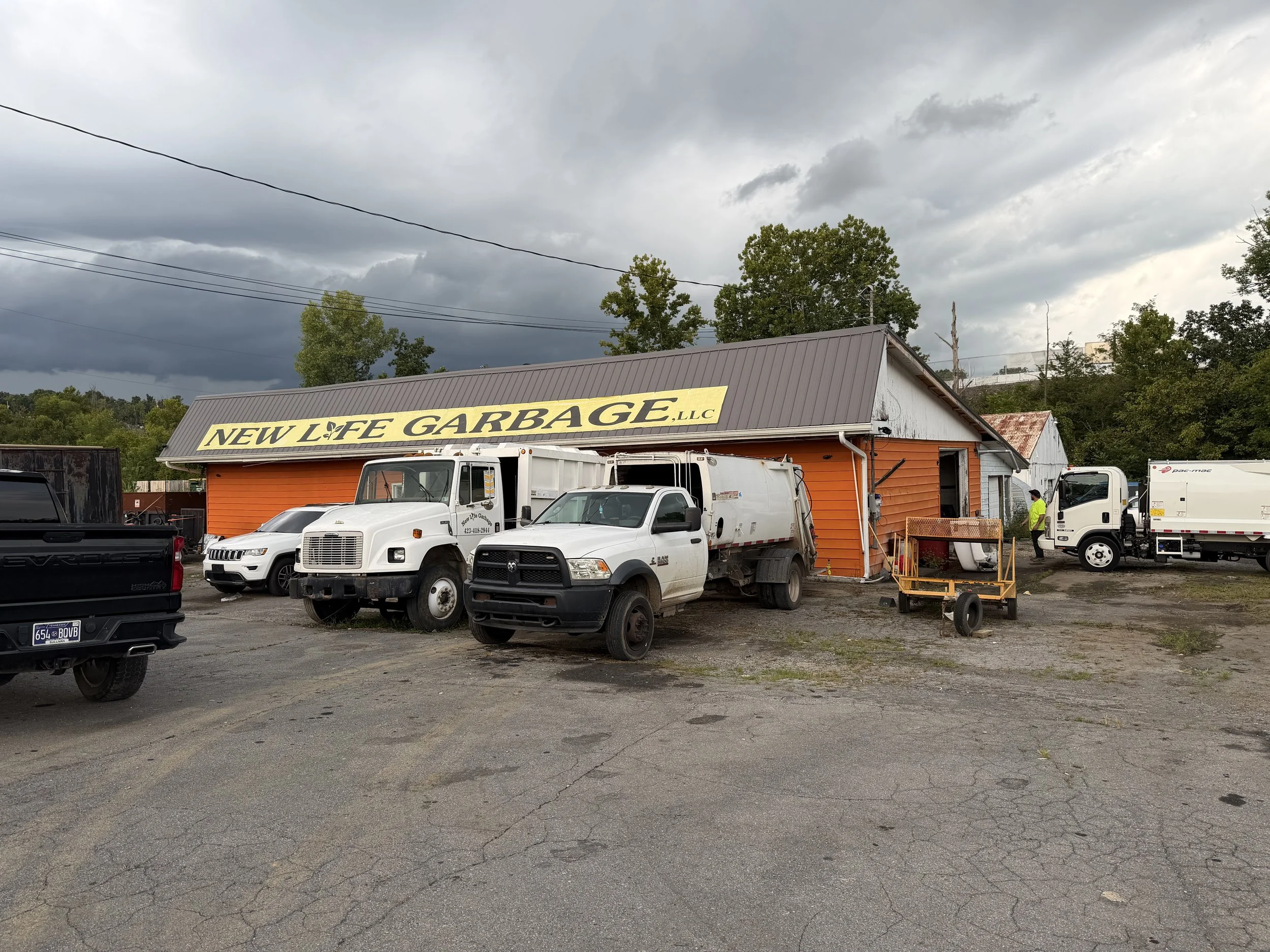 A parking lot with several trucks and cars in front of an orange building with a yellow sign reading 'New Life Garbage, LLC' under a cloudy sky.