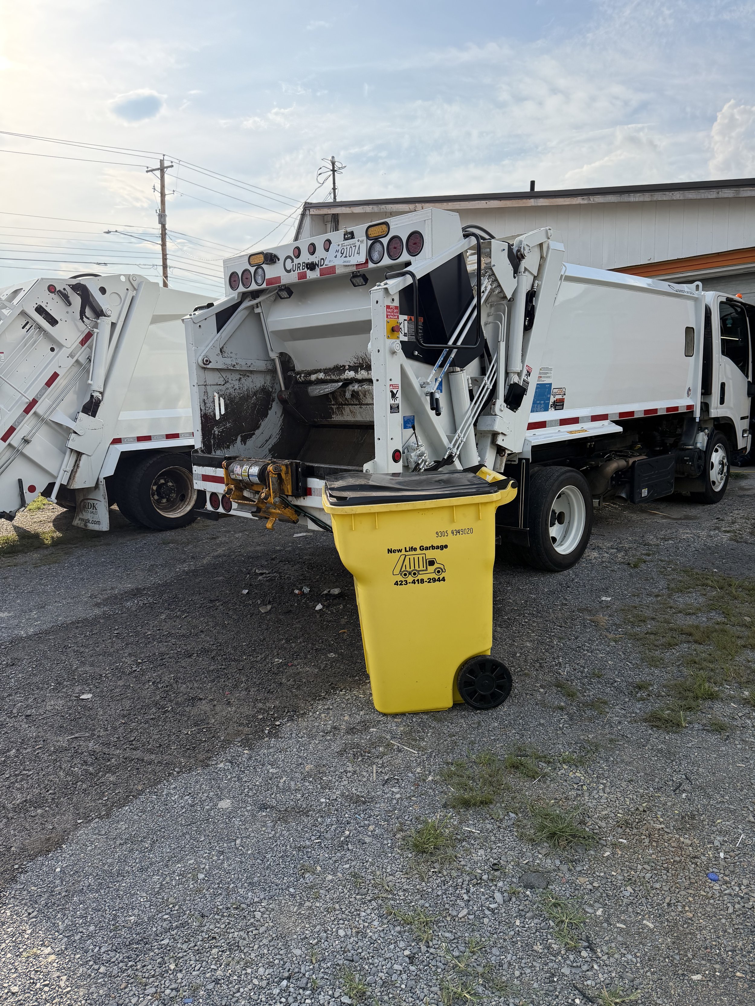 A white garbage truck with hydraulic lifting mechanism parked outdoors under a cloudy sky, with power lines overhead. A yellow trash bin labeled 'New Life Garbage' is in front of the truck.