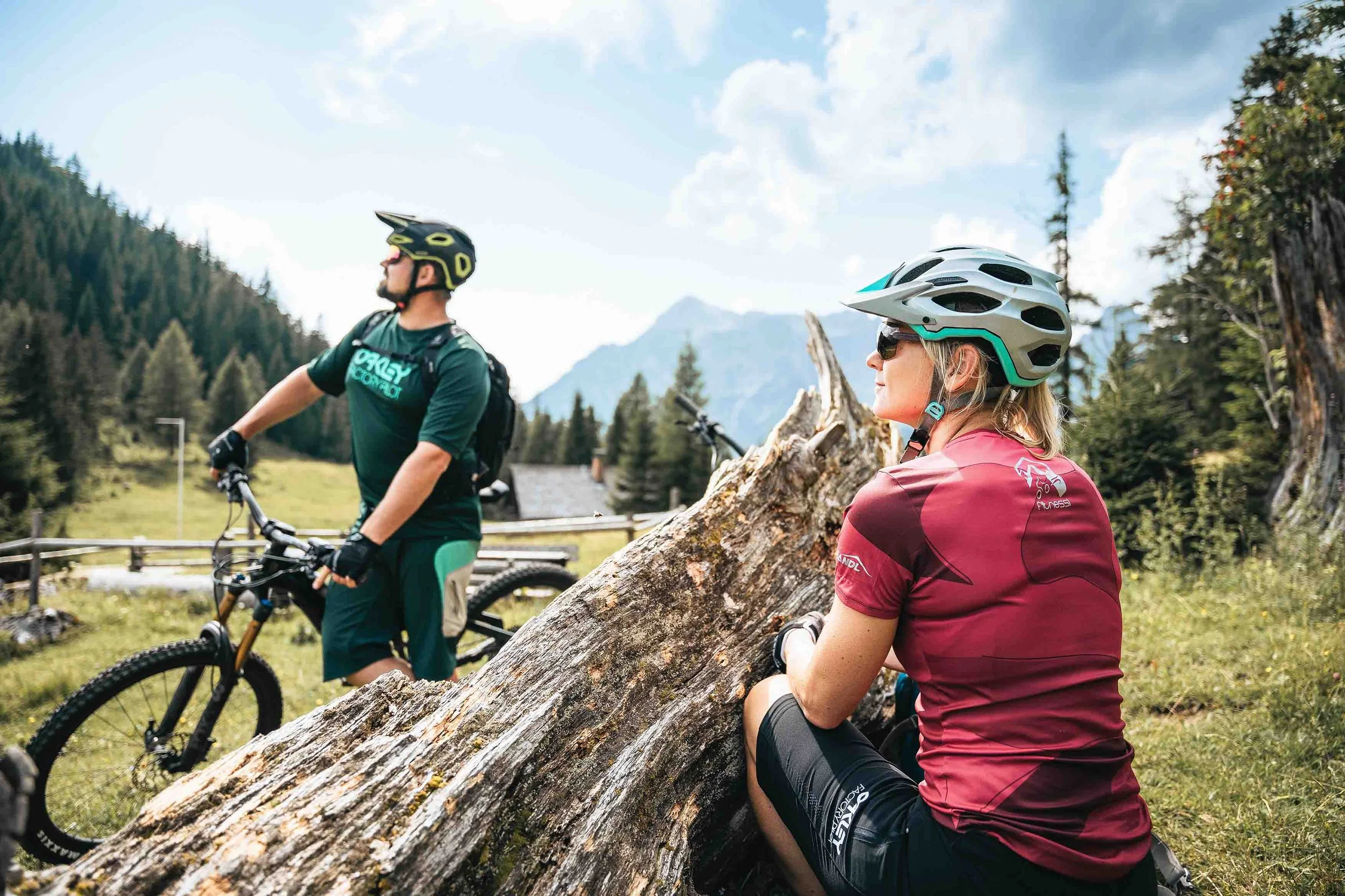 Zwei Radfahrer mit Helmen machen eine Pause im Freien. Eine Frau sitzt auf einem Baumstamm, während ein Mann neben seinem Fahrrad steht. Im Hintergrund sind Berge und ein Wald zu sehen.
