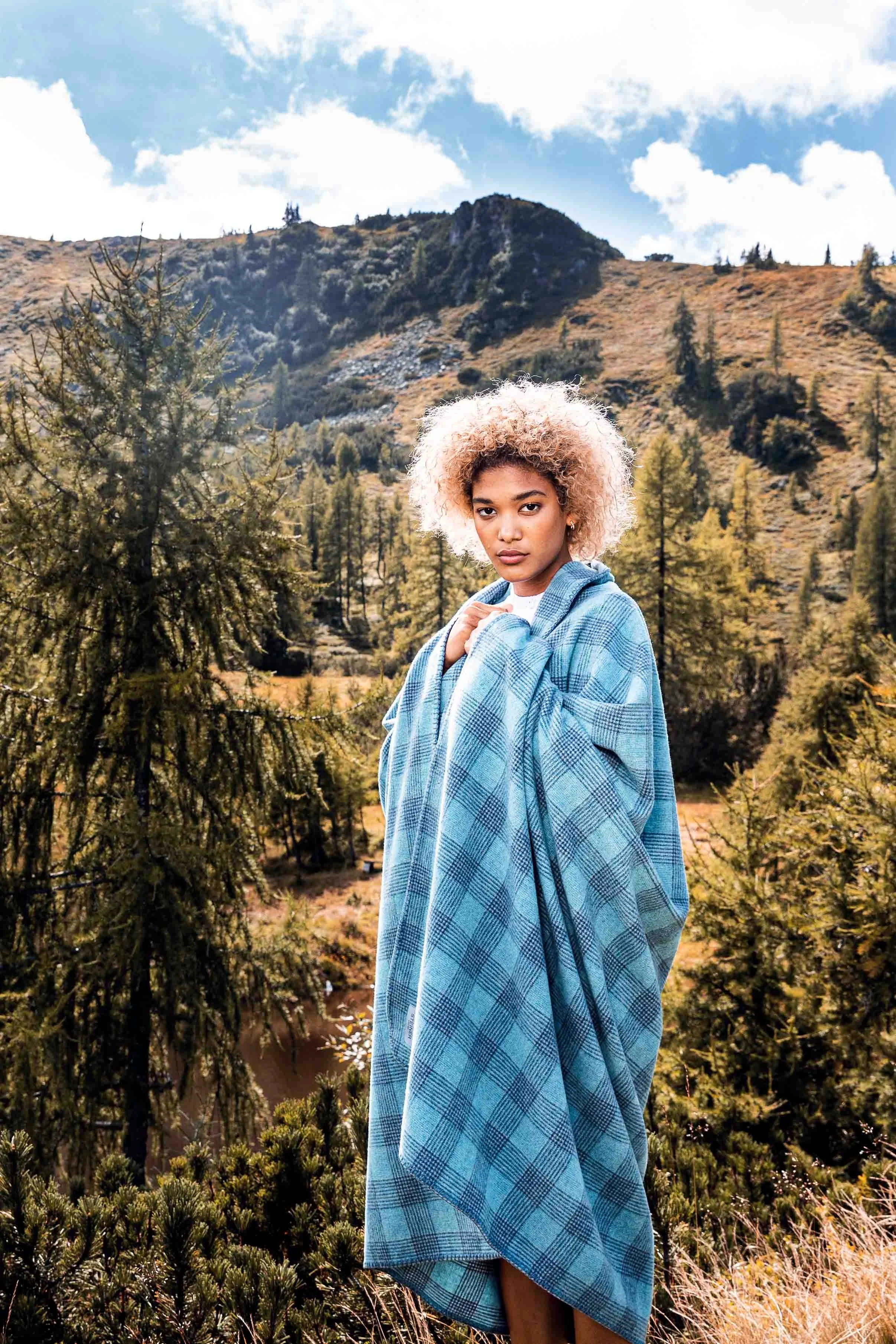 Frau mit blauer Decke steht in einer malerischen Berglandschaft mit Bäumen und hügeligem Terrain.