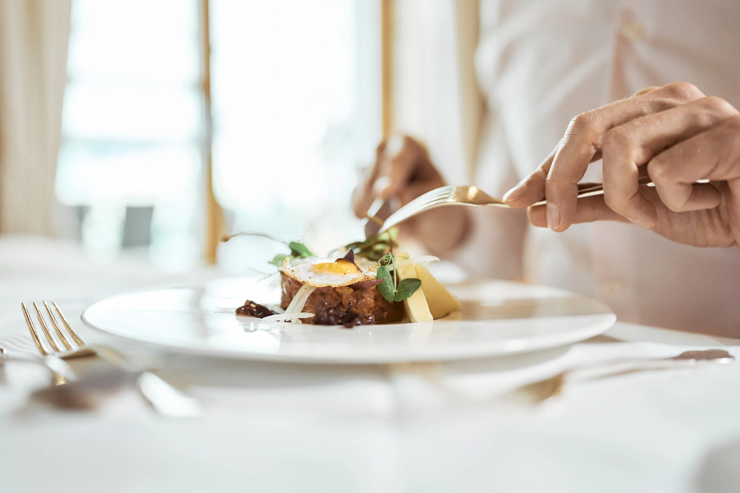 Person isst ein Gourmetgericht mit Besteck in einem eleganten Restaurant, mit fokus auf einem kunstvoll angerichteten Teller.