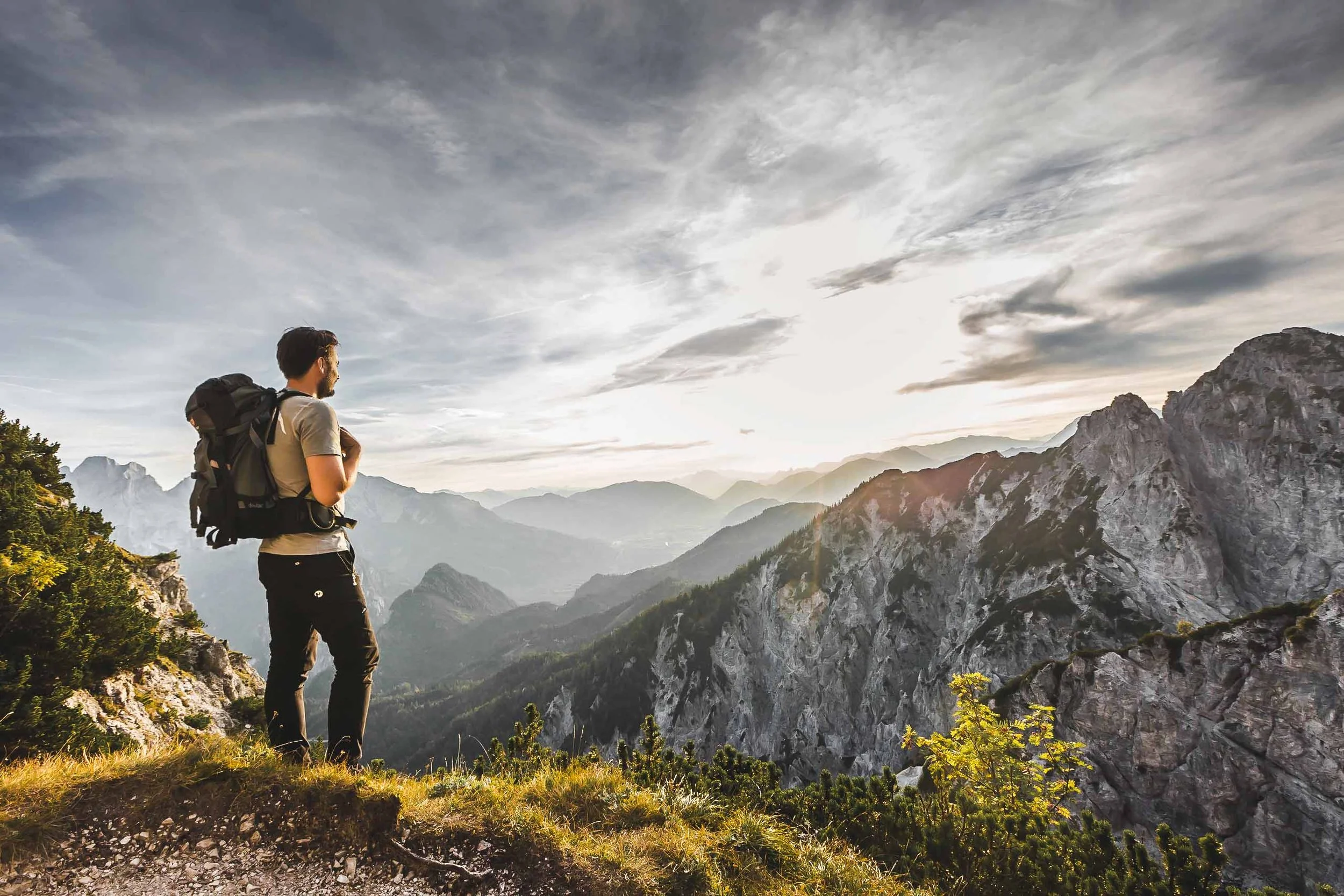 Ein Wanderer mit Rucksack steht auf einem Bergpfad und blickt auf eine beeindruckende Berglandschaft unter einem bewölkten Himmel.