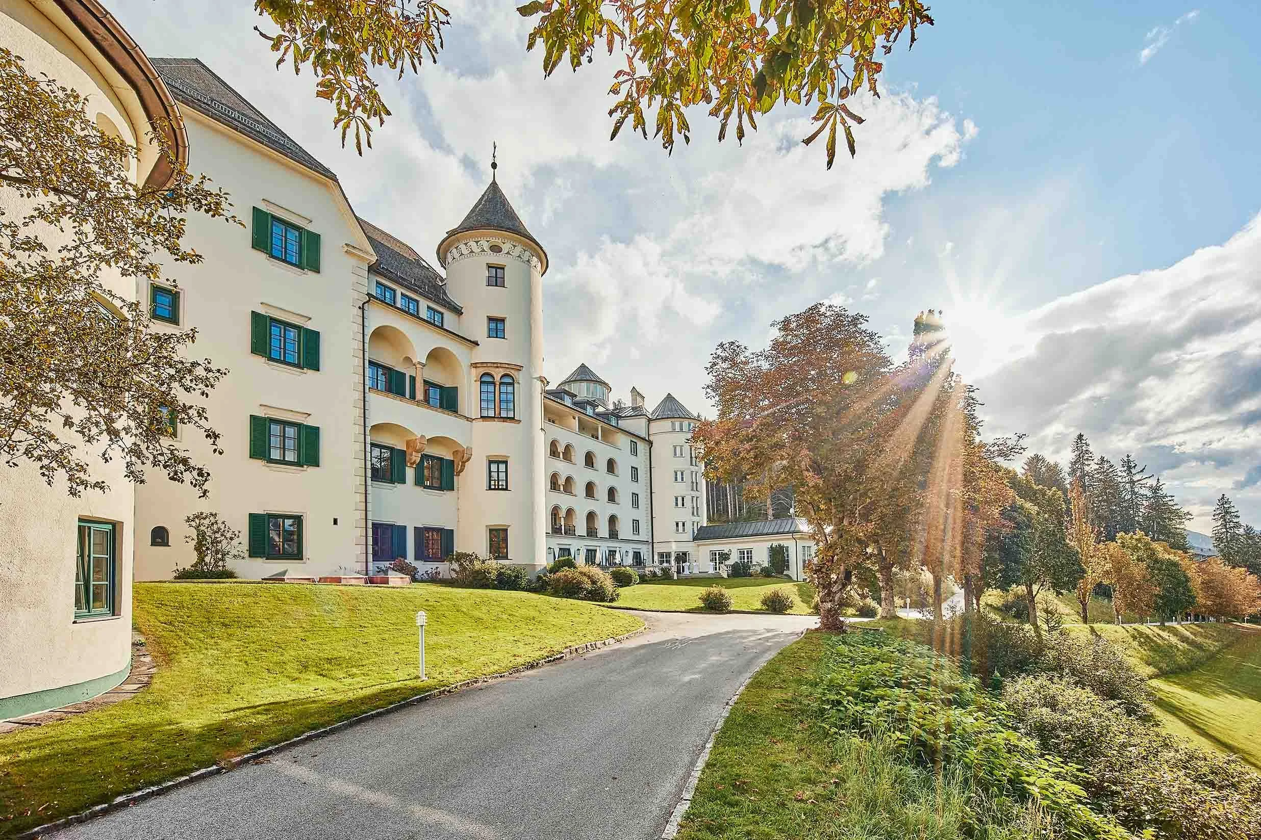 Schlossartiges Gebäude mit Türmen und grünen Fensterläden, umgeben von Bäumen und gepflegtem Rasen, bei sonnigem Wetter.