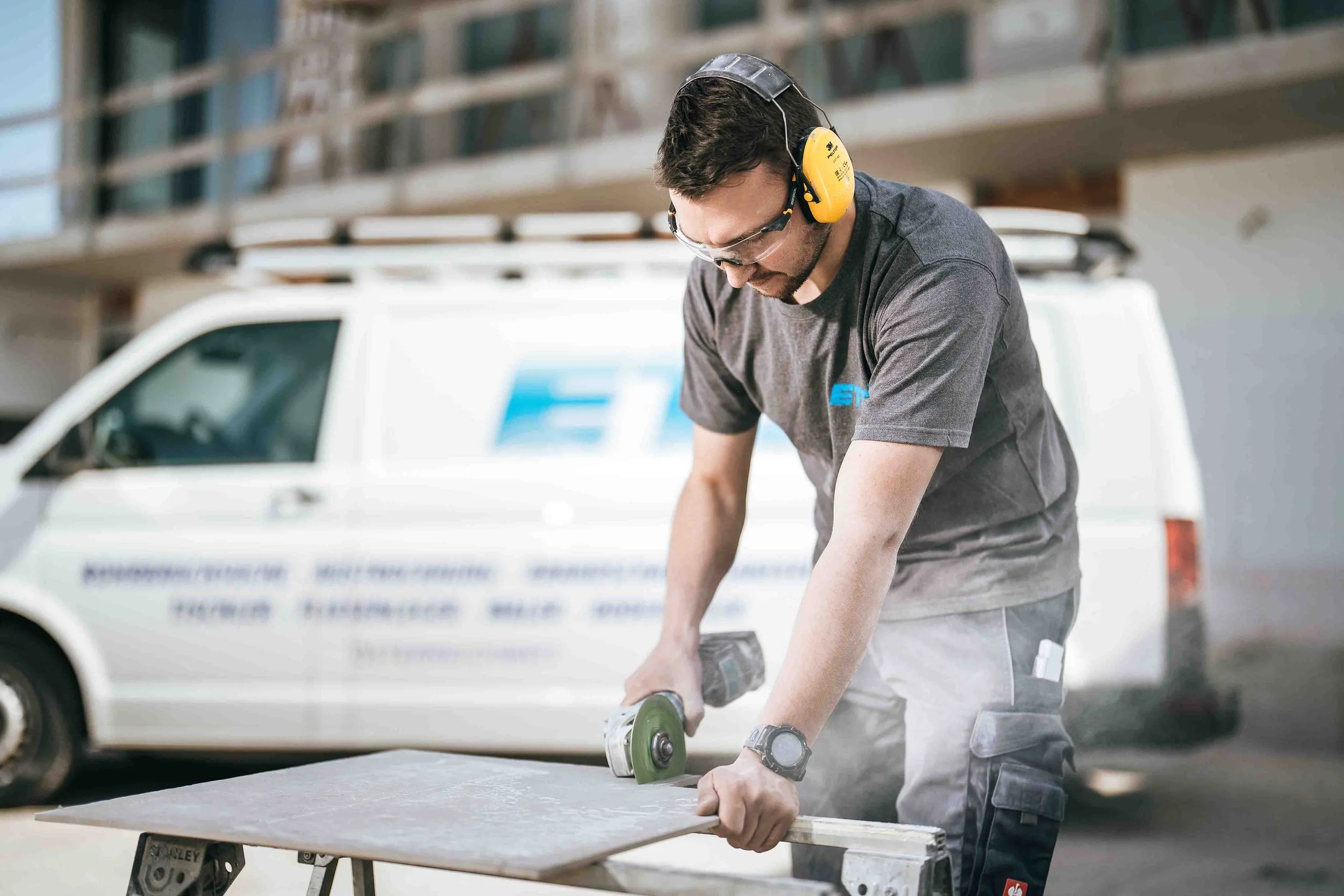 Ein Mitarbeiter von ETS trägt Gehörschutz und Schutzbrille, während er mit einem Winkelschleifer auf einer Baustelle arbeitet. Im Hintergrund steht ein weißer Lieferwagen.