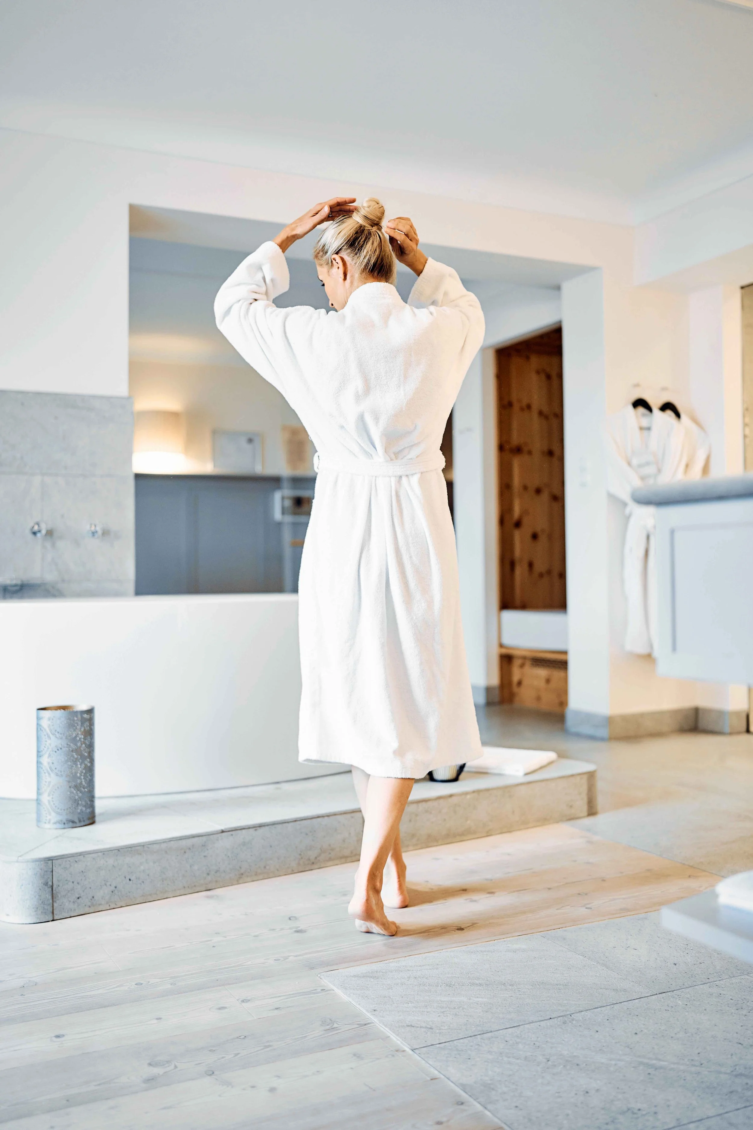 Person in einem weißen Bademantel, die sich im Badezimmer die Haare hochsteckt. Moderner Raum mit Badewanne und Hölzboden.