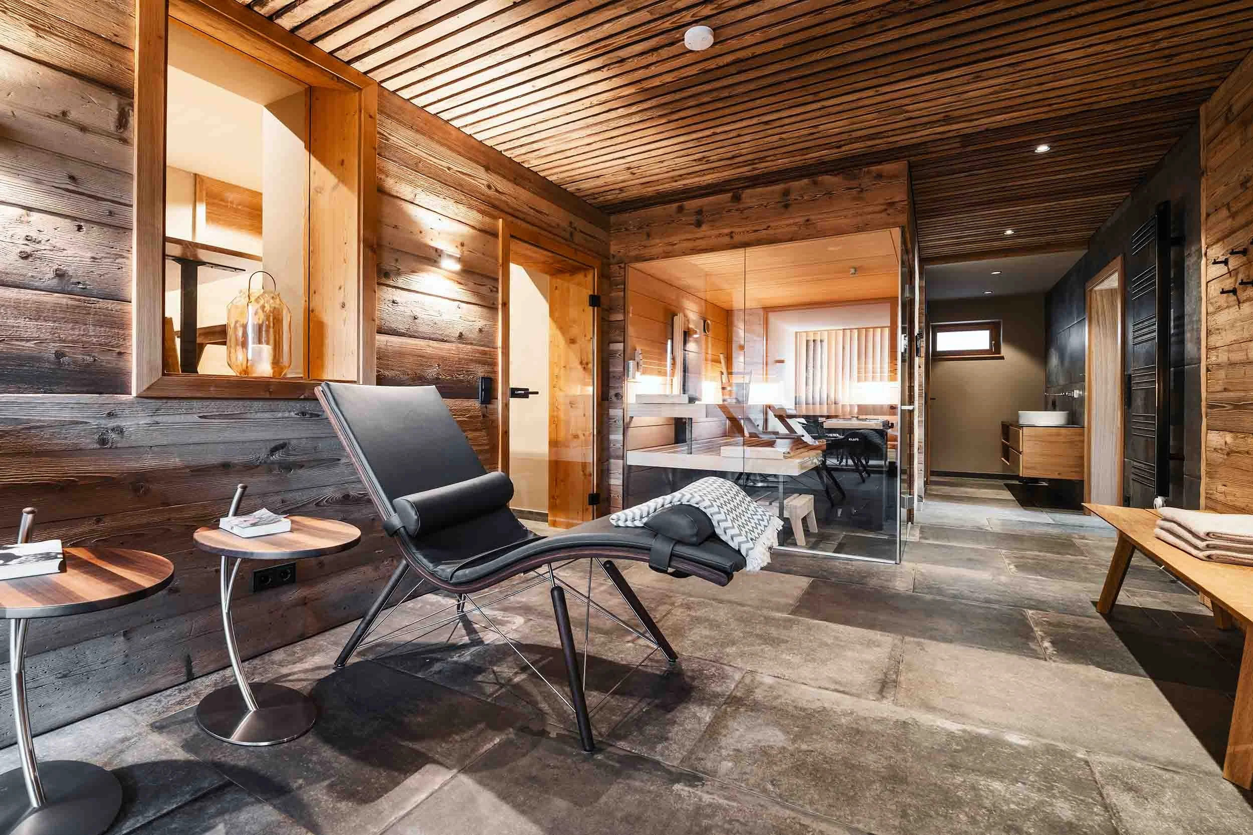 Eine gemütliche, moderne Wellnessoase aus Holz. Es gibt eine Liege mit Kissen, zwei kleine Beistelltische, Handtücher auf einer Holzbank und einen Glaskubus, der eine Sauna oder einen Ruheraum andeutet. Die Beleuchtung ist warm, und die Wände sind au
