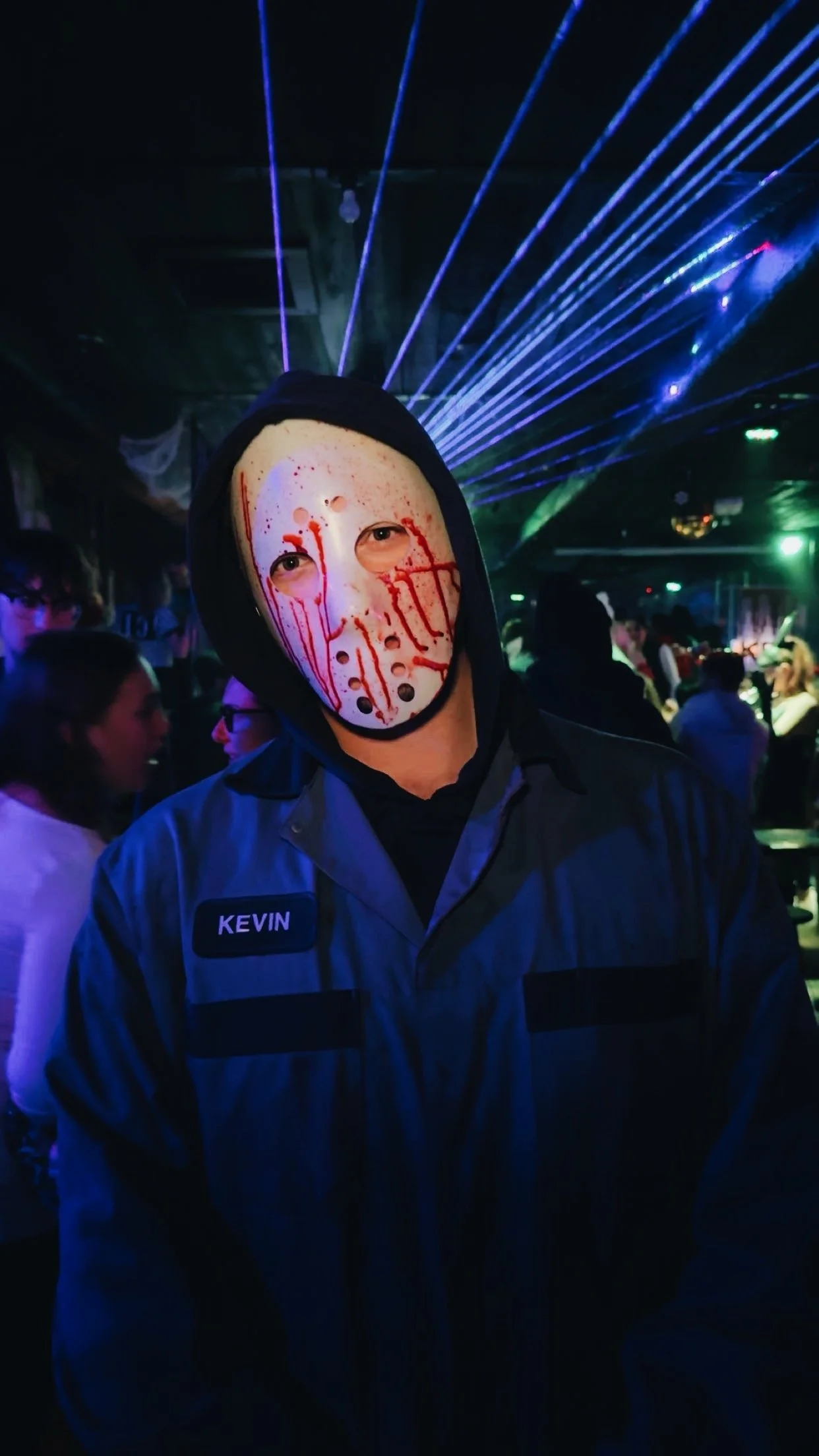 Person dressed as Ghostface from 'Scream' wearing a white mask with red blood-like markings, black hoodie, and dark jacket in a nightclub with colorful laser lights and other people in the background.