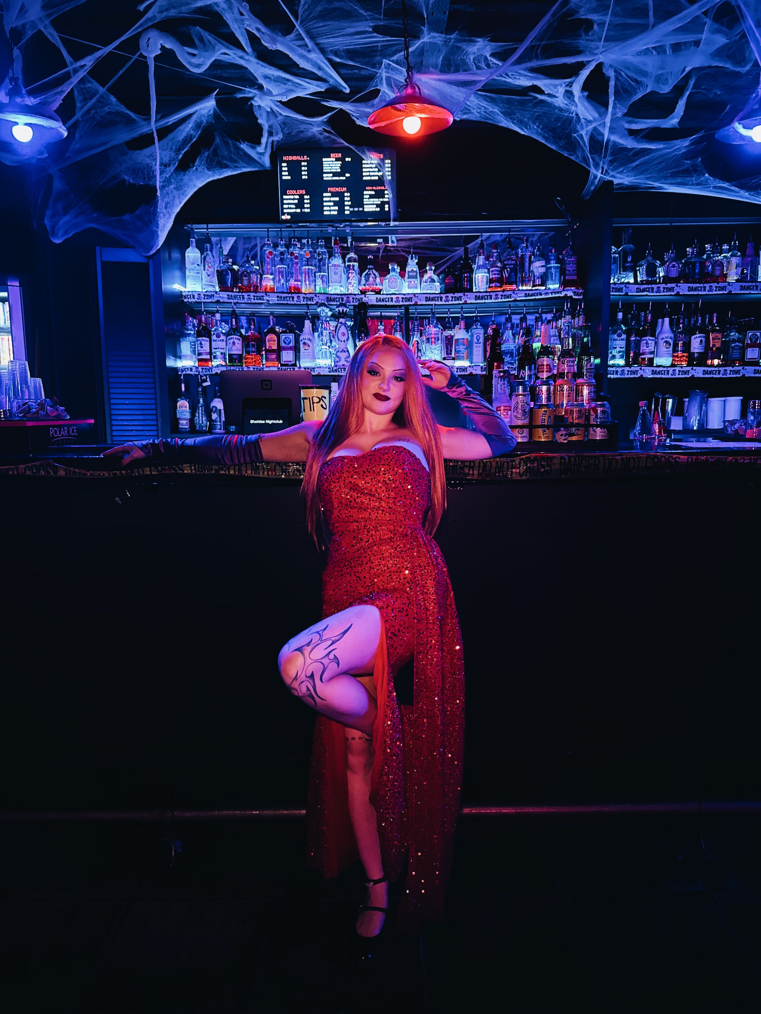 A woman with long red hair and black lipstick posing in a red, sparkly dress with a high slit in a dimly lit bar decorated for Halloween, with spider web decorations on the ceiling and various bottles of alcohol behind the bar.