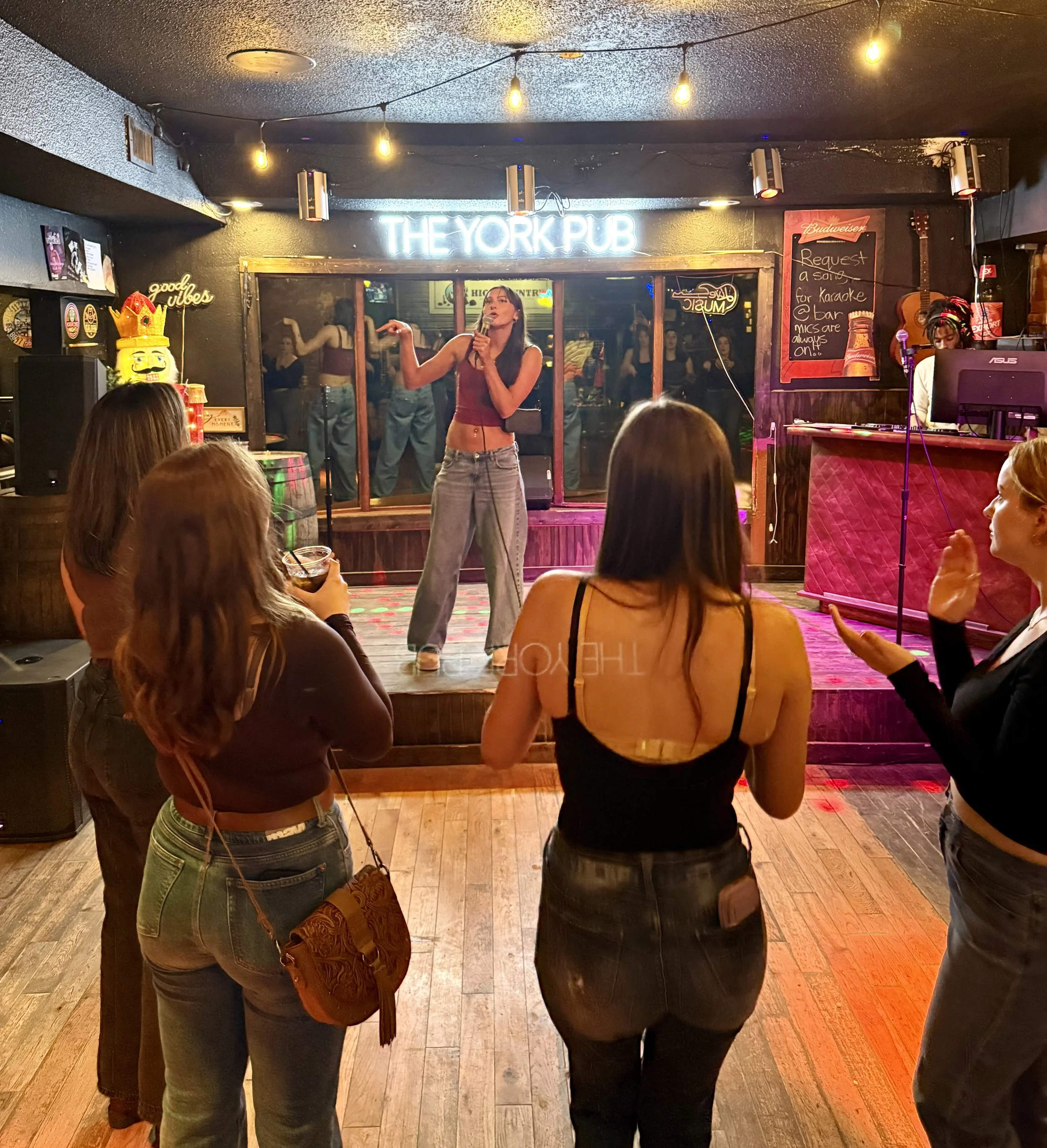 A woman performing on stage at The York Pub with an audience watching. The stage has a neon sign overhead and a DJ in the corner.