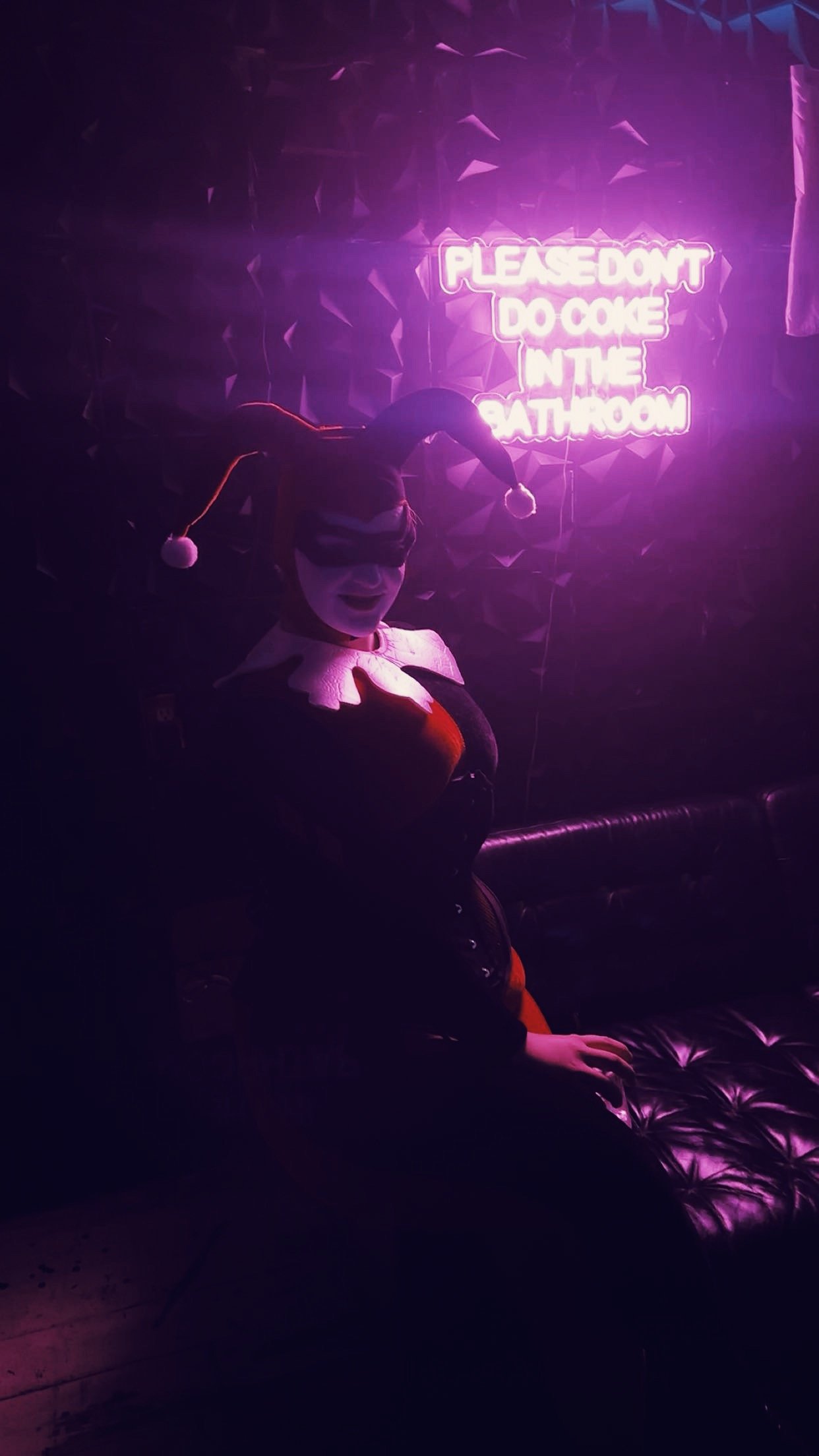 A person dressed as a clown with a hat and makeup, sitting on a dark, tufted bench in front of a neon pink sign that reads 'PLEASE DON'T DO COKE IN THE BATHROOM' in a dimly lit room with purple lighting.