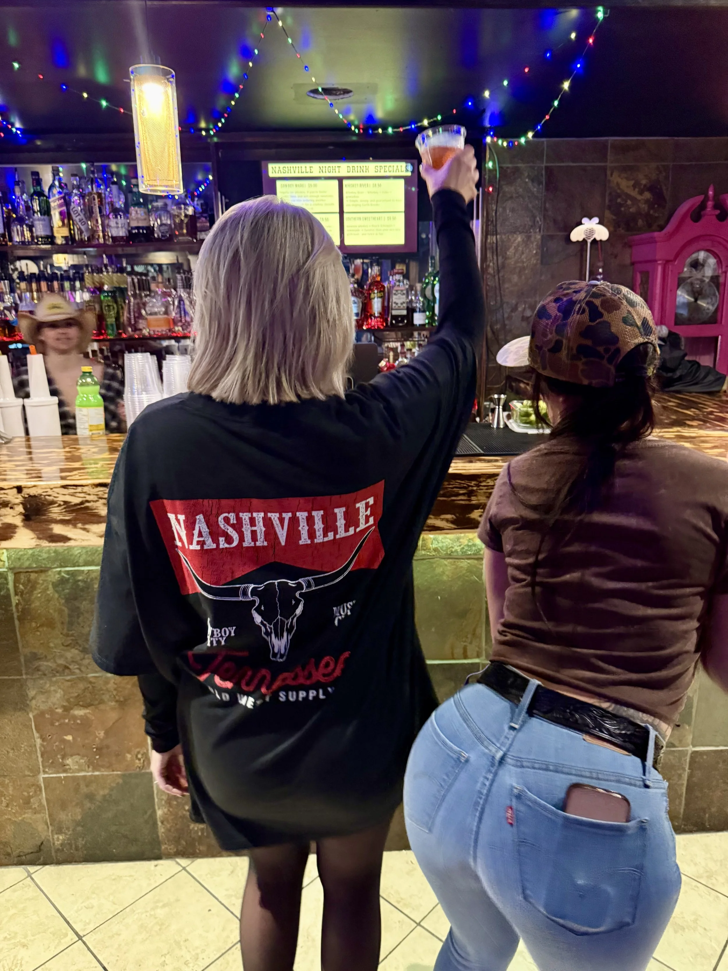 Two women at a bar, one is raising a drink in a toast, another is leaning on the bar, wearing a camouflage cap and blue jeans.