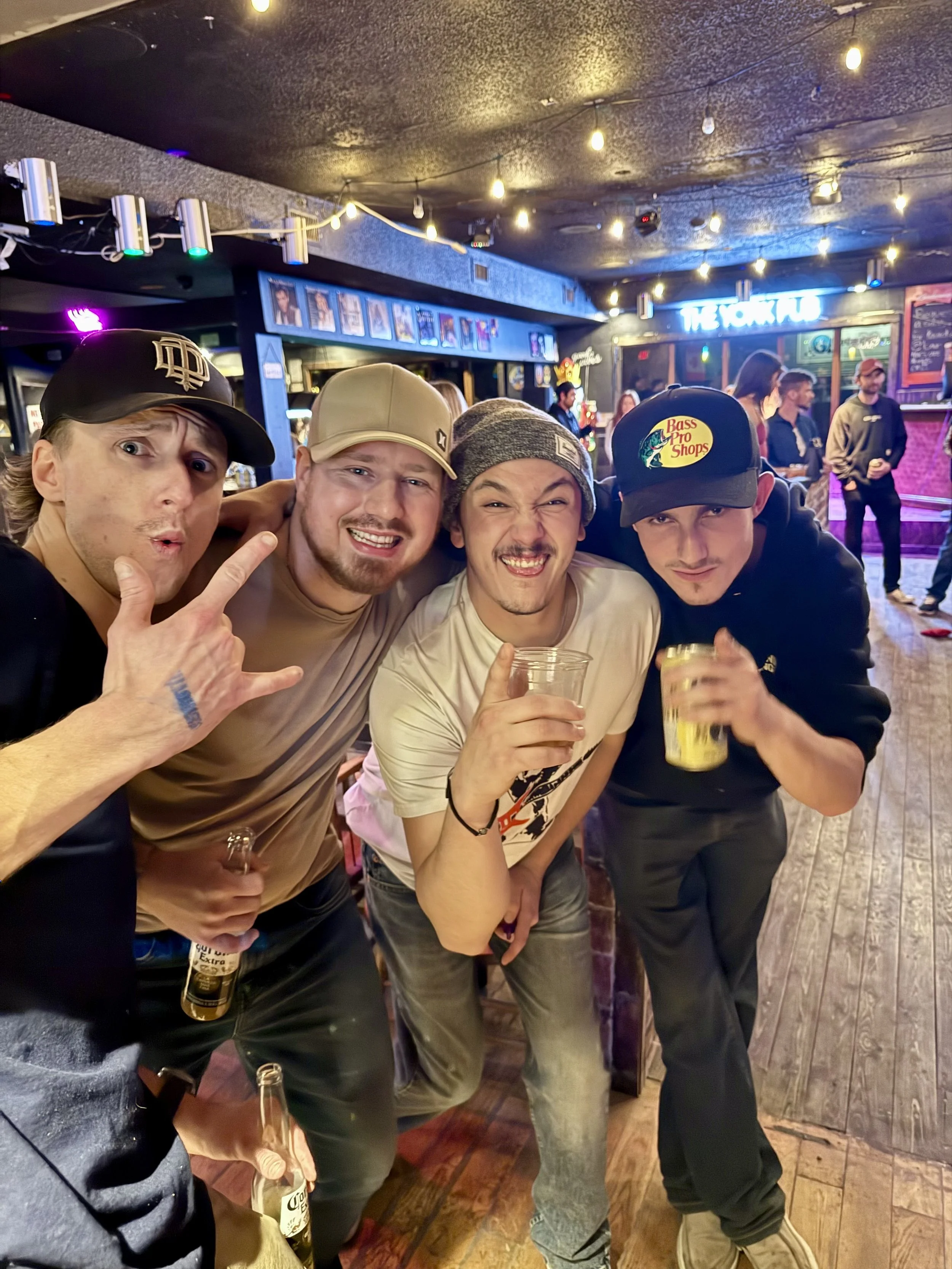 Four young men at a bar, smiling and holding drinks, with other patrons and a neon sign in the background.
