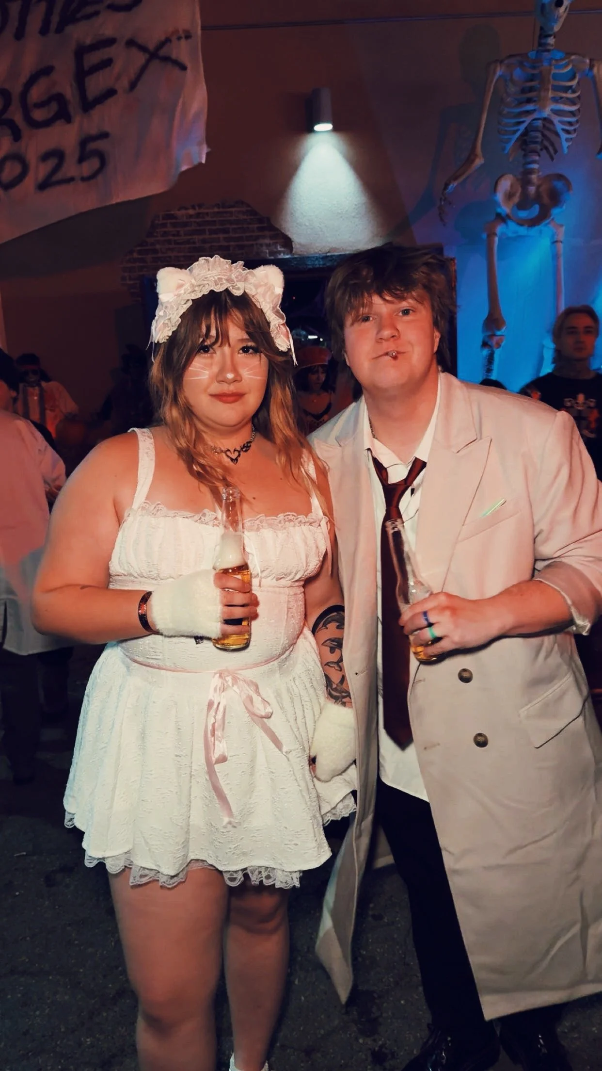 Two people in costume at a party, with a woman dressed as a cat and a man in a white suit holding beer bottles, in a dimly lit room with skeleton decorations.
