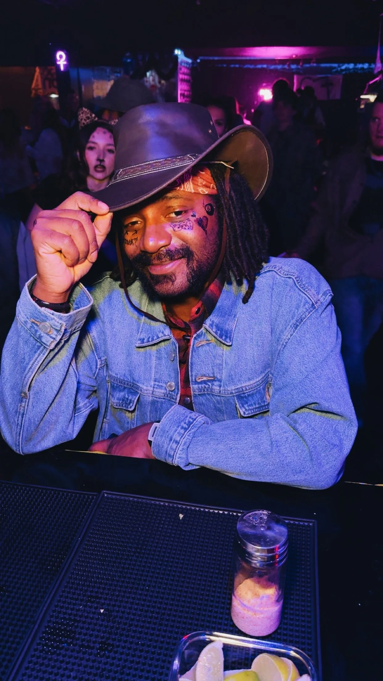A man with face tattoos, wearing a denim jacket and a wide-brimmed hat, sitting at a bar in a dimly lit, colorful club, smiling and adjusting his hat.