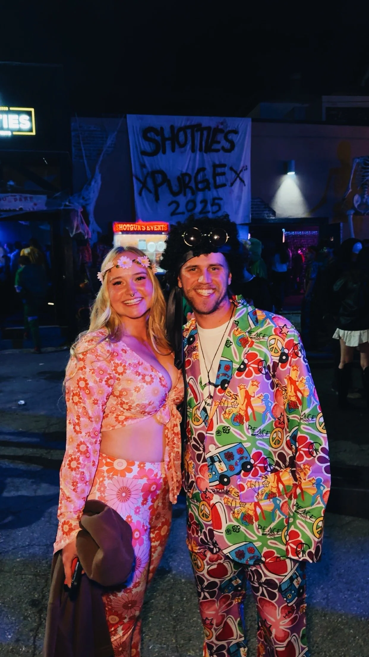 A woman and a man dressed in colorful, 1960s-style outfits at a party with a banner that reads 'SHOTTIES XPURGE X 2025' in the background.