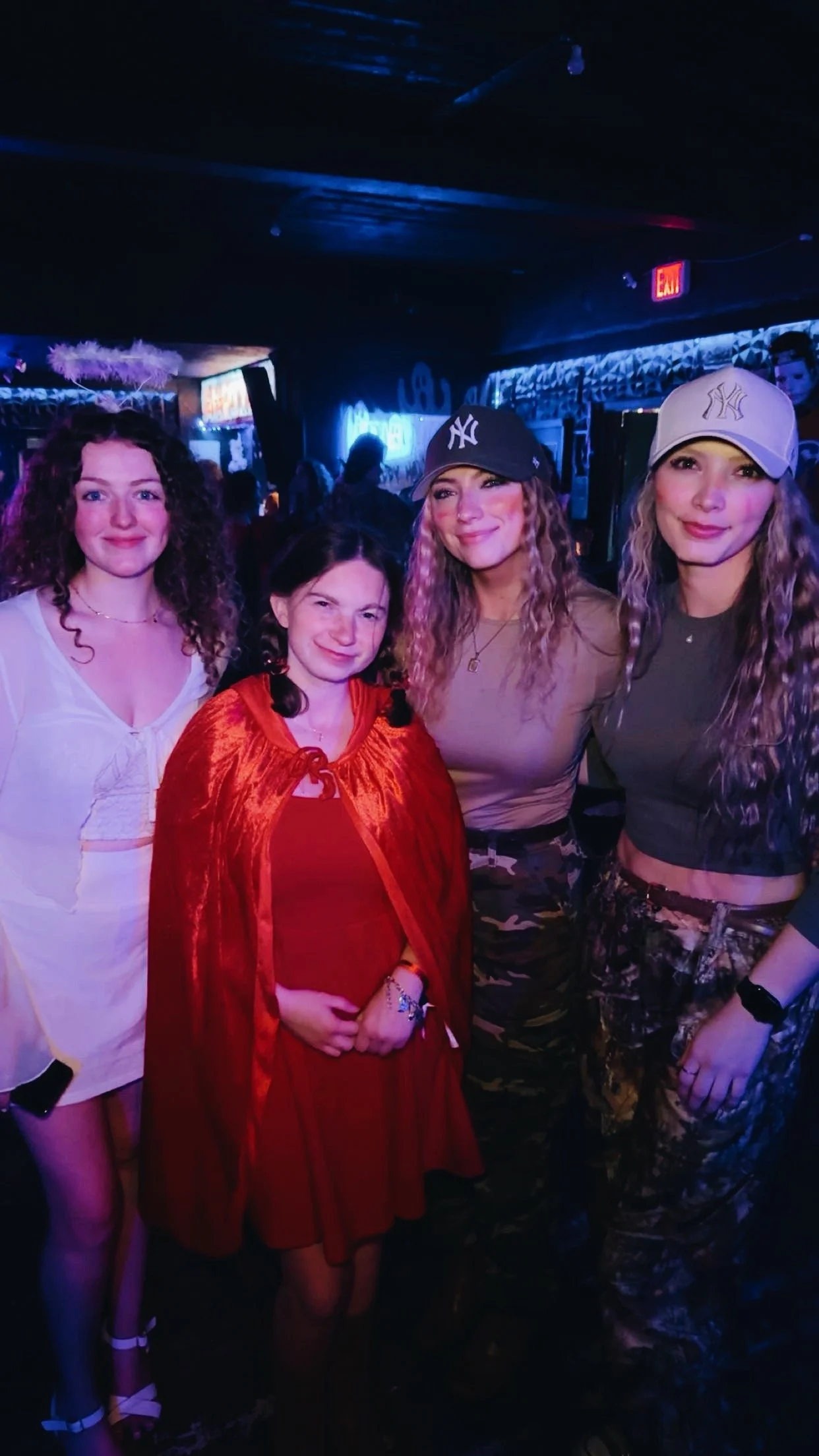 Four young women standing together in a dimly lit nightclub, smiling for the camera, with neon signs visible in the background.