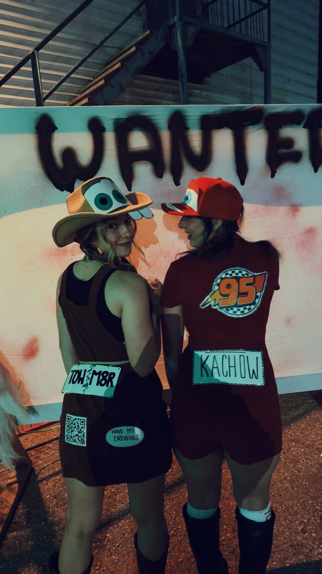 Two women dressed as racers at a race-themed event, wearing colorful hats with cartoonish eyes, standing in front of a large 'WANTED' sign, with one woman in a black outfit with a name tag 'Toni M8R' and the other in a red outfit with a name tag 'Kac
