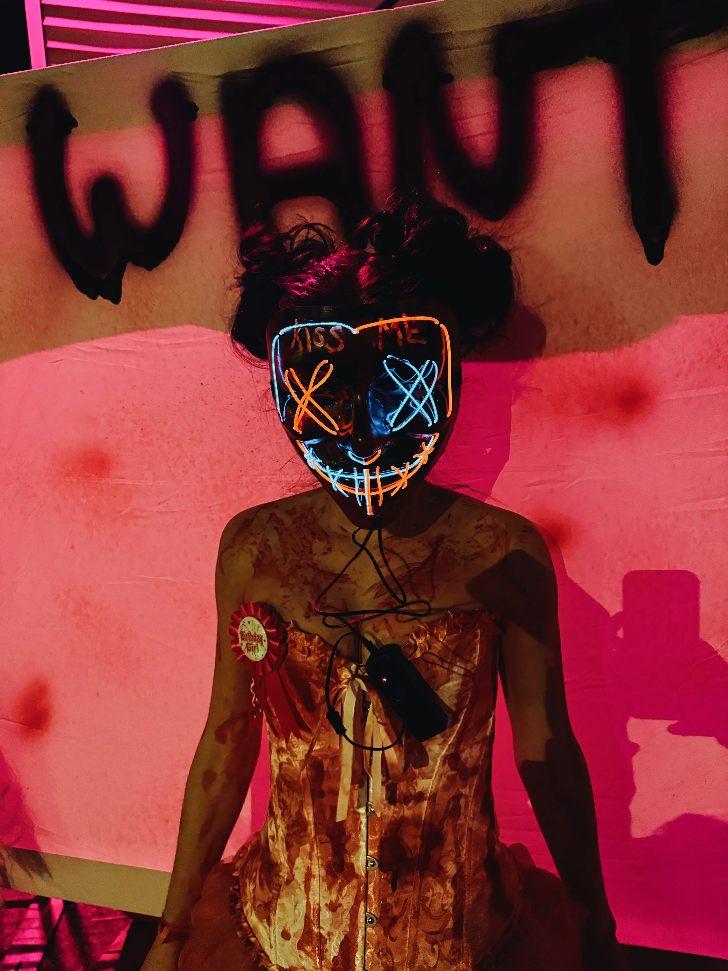 Person wearing a glowing mask with a smiling face design, the words 'KISS ME' on the forehead, and the person is dressed in a satin dress with a birthday badge. The background features pink lighting and dark graffiti writing.