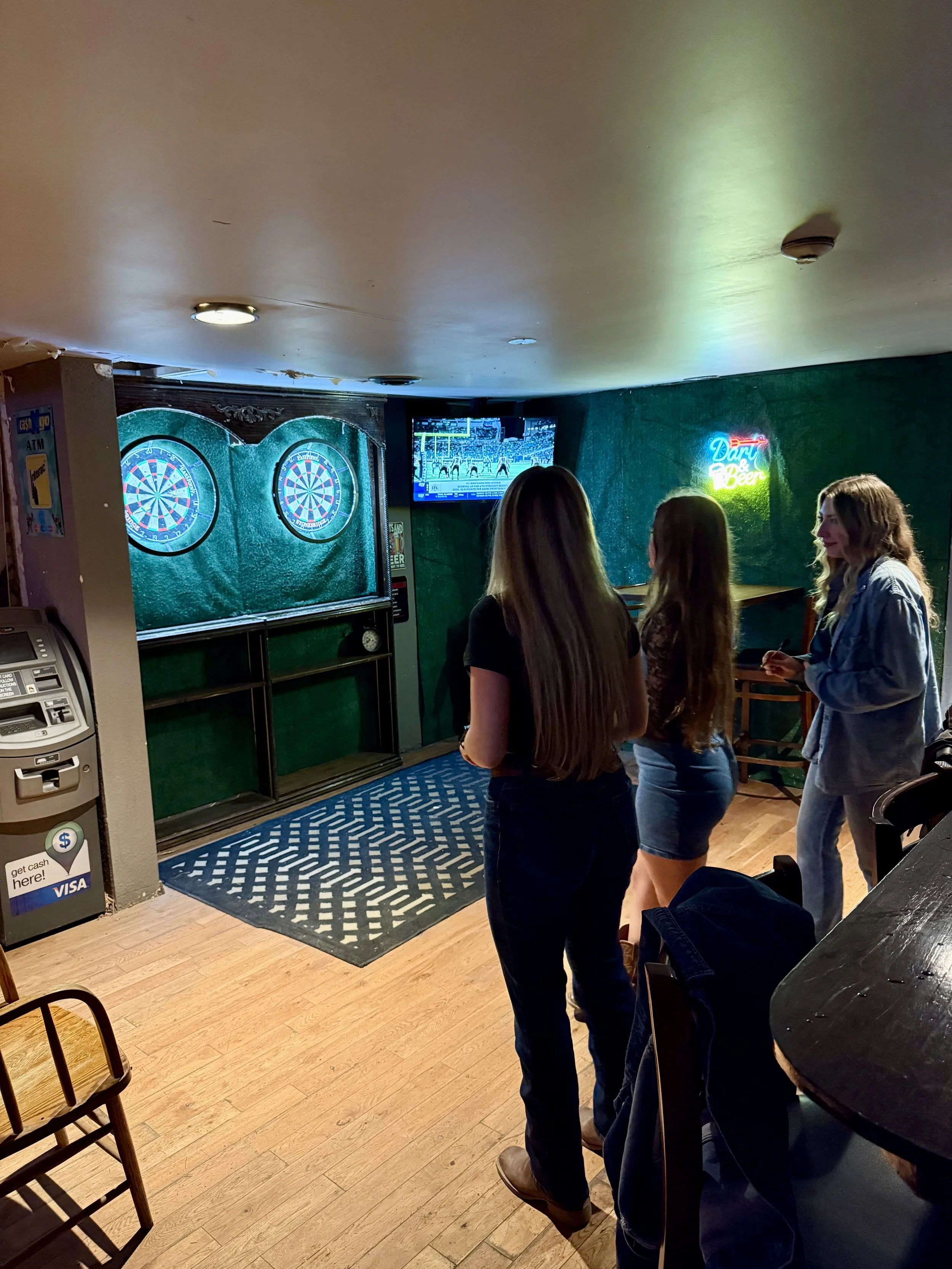 Three women stand in a bar, watching two dartboards and a TV screen showing a sports game. There is a neon sign on the wall for dark beer, a credit card ATM machine on the side, and a patterned rug on the wooden floor.