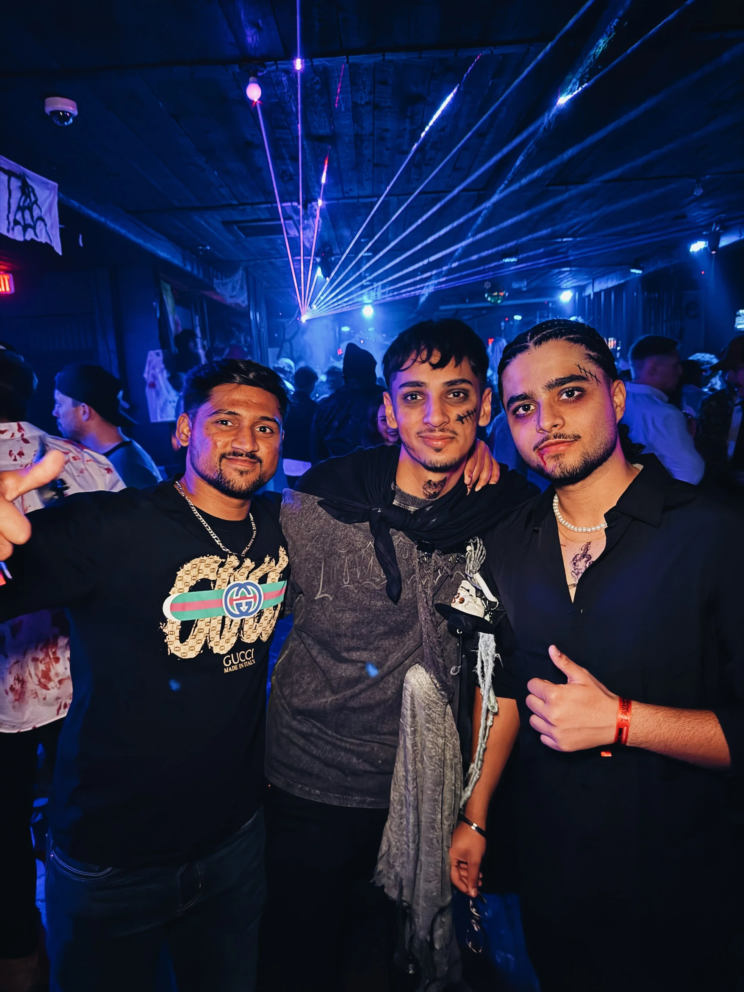 Three young men posing together in a nightclub with blue and purple laser lights and fog in the background.