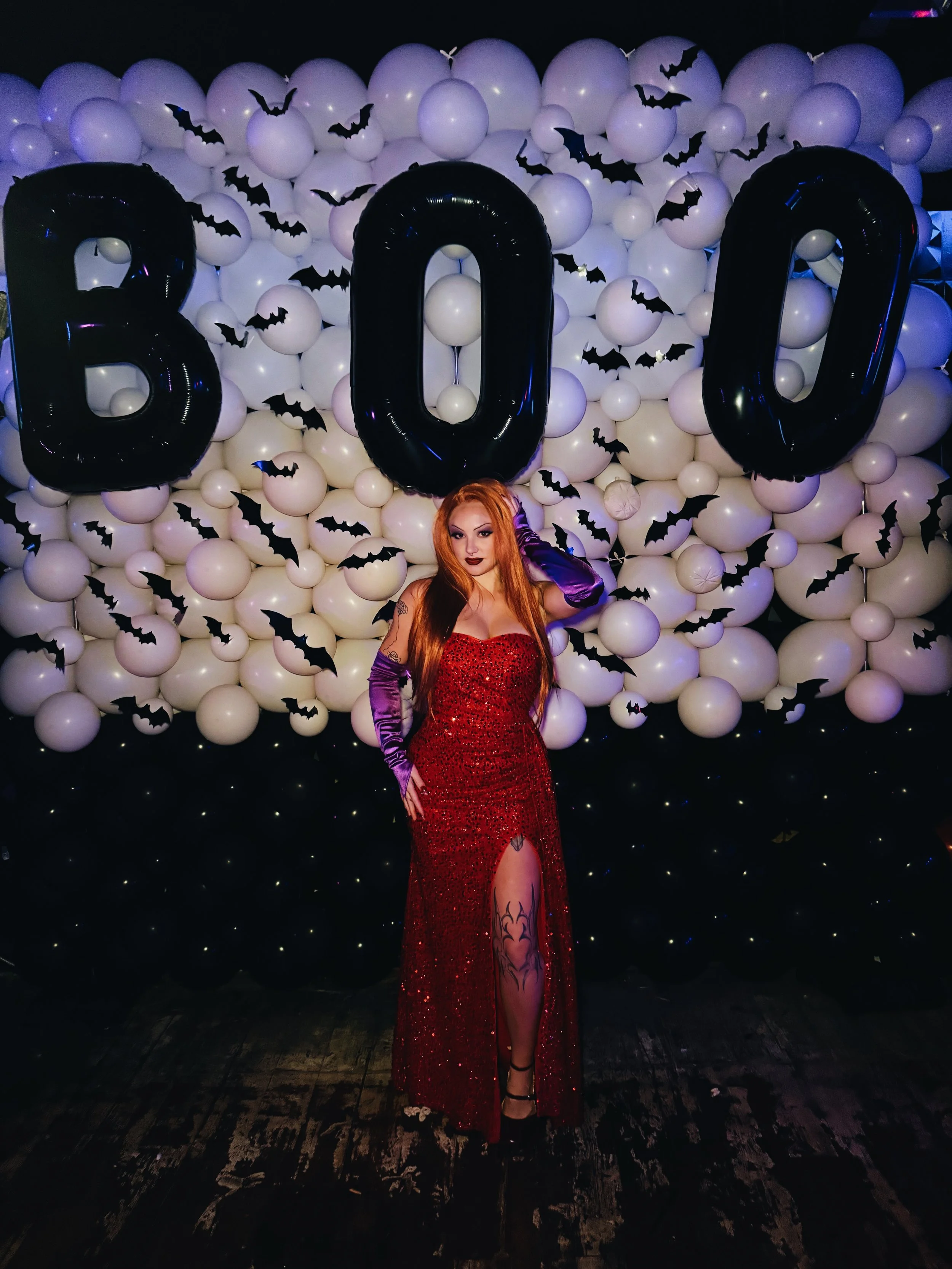 A woman with long red hair wearing a red sparkly dress with a high slit and black heels, standing in front of a Halloween-themed backdrop with large black balloons spelling 'BOO', white balloons, and black paper bats, at a party or event.
