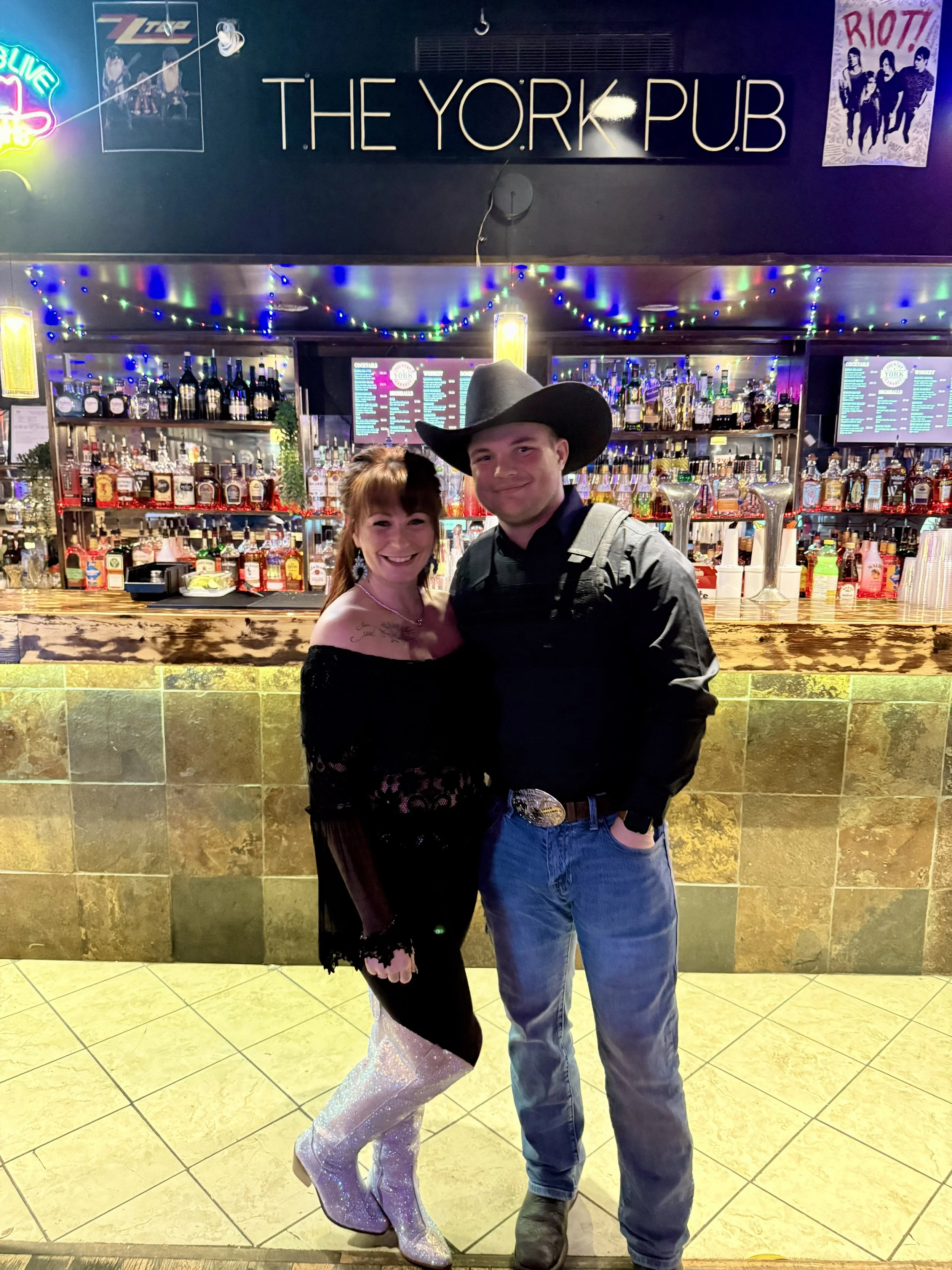 A woman and man dressed in country western attire standing together in front of a bar at The York Pub. The woman wears a black off-shoulder top with lace details, shiny silver boots, and has a tattoo on her shoulder. The man wears a black cowboy hat,