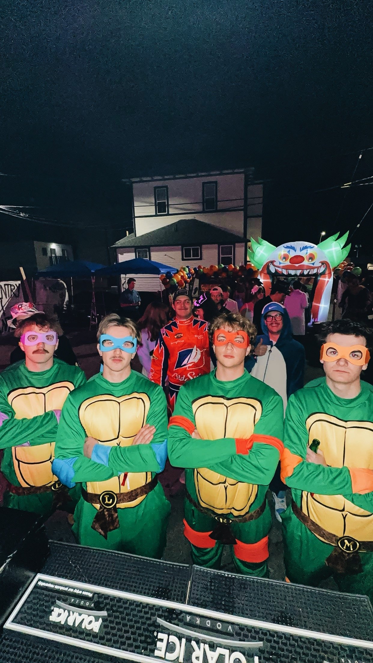 People dressed in Teenage Mutant Ninja Turtles costumes at a nighttime outdoor event with a clown-themed inflatable slide in the background.