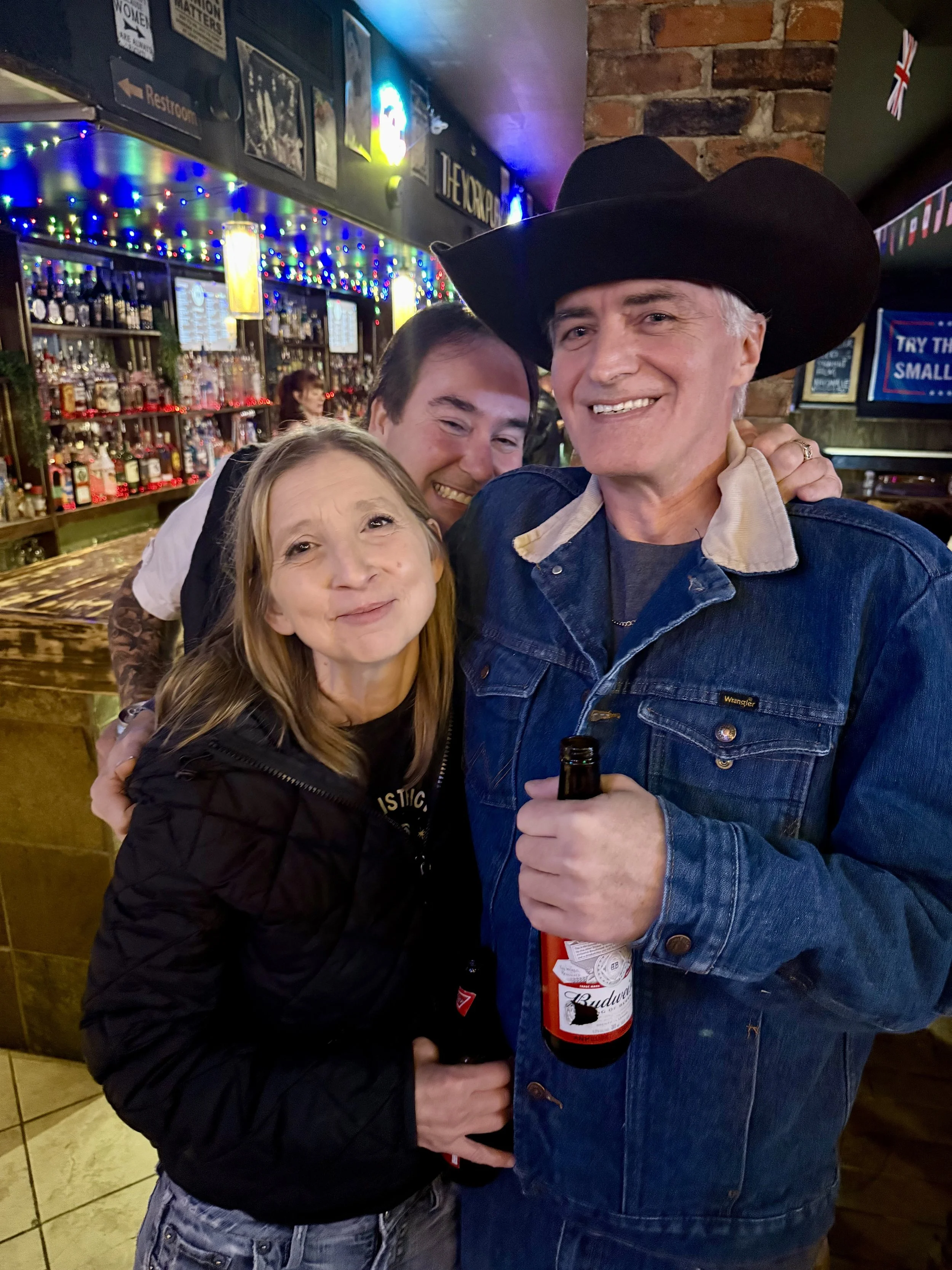 Three people smiling and posing together in a bar. The man on the right is wearing a cowboy hat and a denim jacket, holding a beer. The woman on the left has blonde hair and is wearing a black jacket. The man in the middle is standing behind them, sm